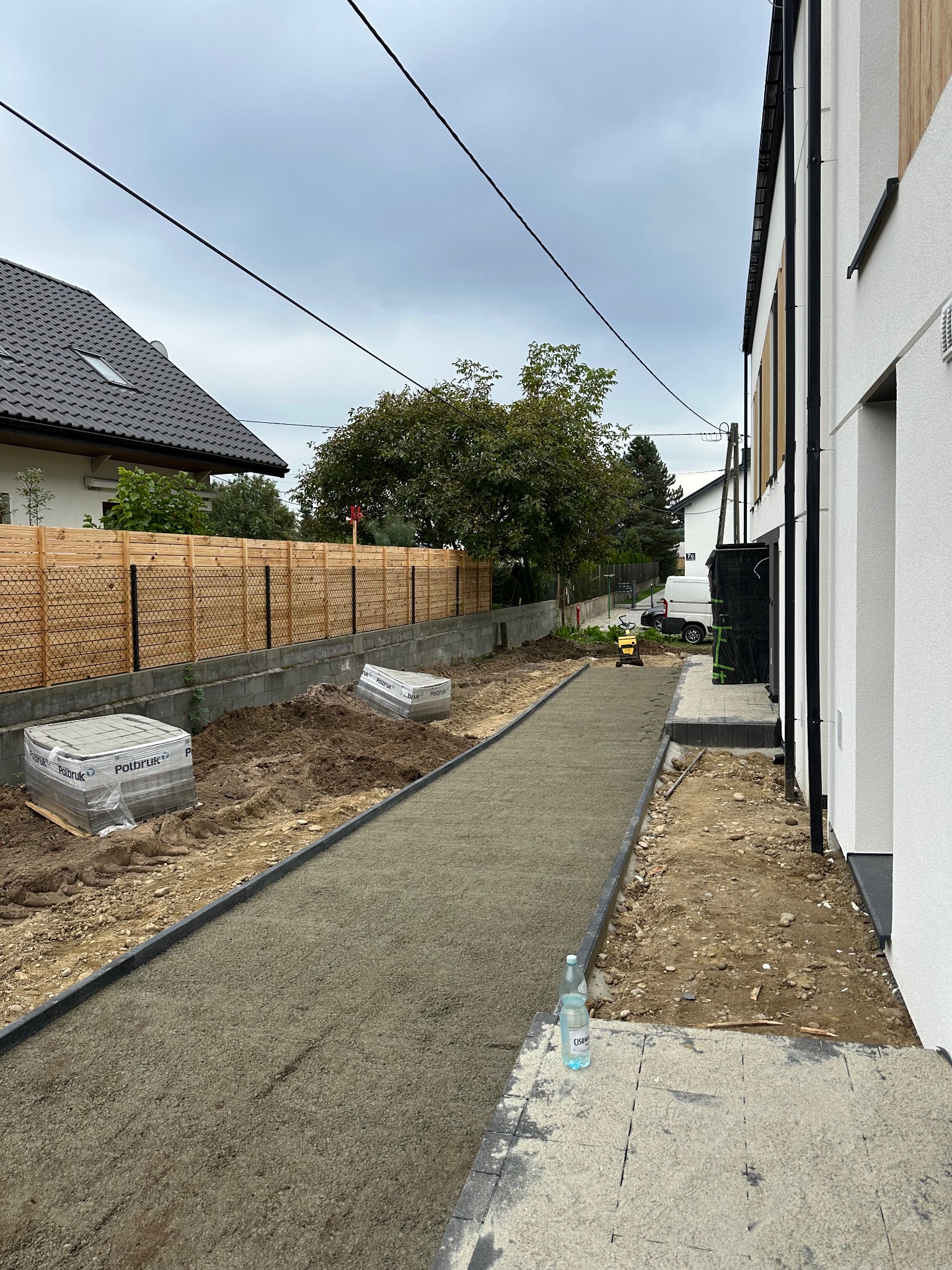 Przygotowanie podłoża pod kostkę brukową na posesji: podsypka wyrównana, obrzeża ułożone, palety kostki czekają na montaż. Widoczny zagęszczarka i butelka wody.