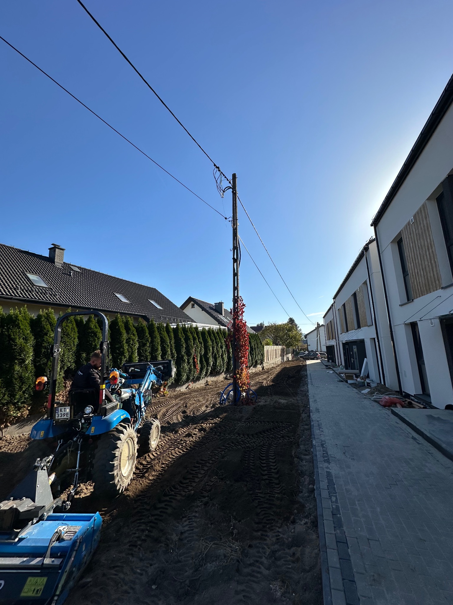 Niebieski traktor przygotowuje teren pod trawnik między nowymi domami a żywopłotem, w tle błękitne niebo i słup energetyczny z pnącą rośliną.