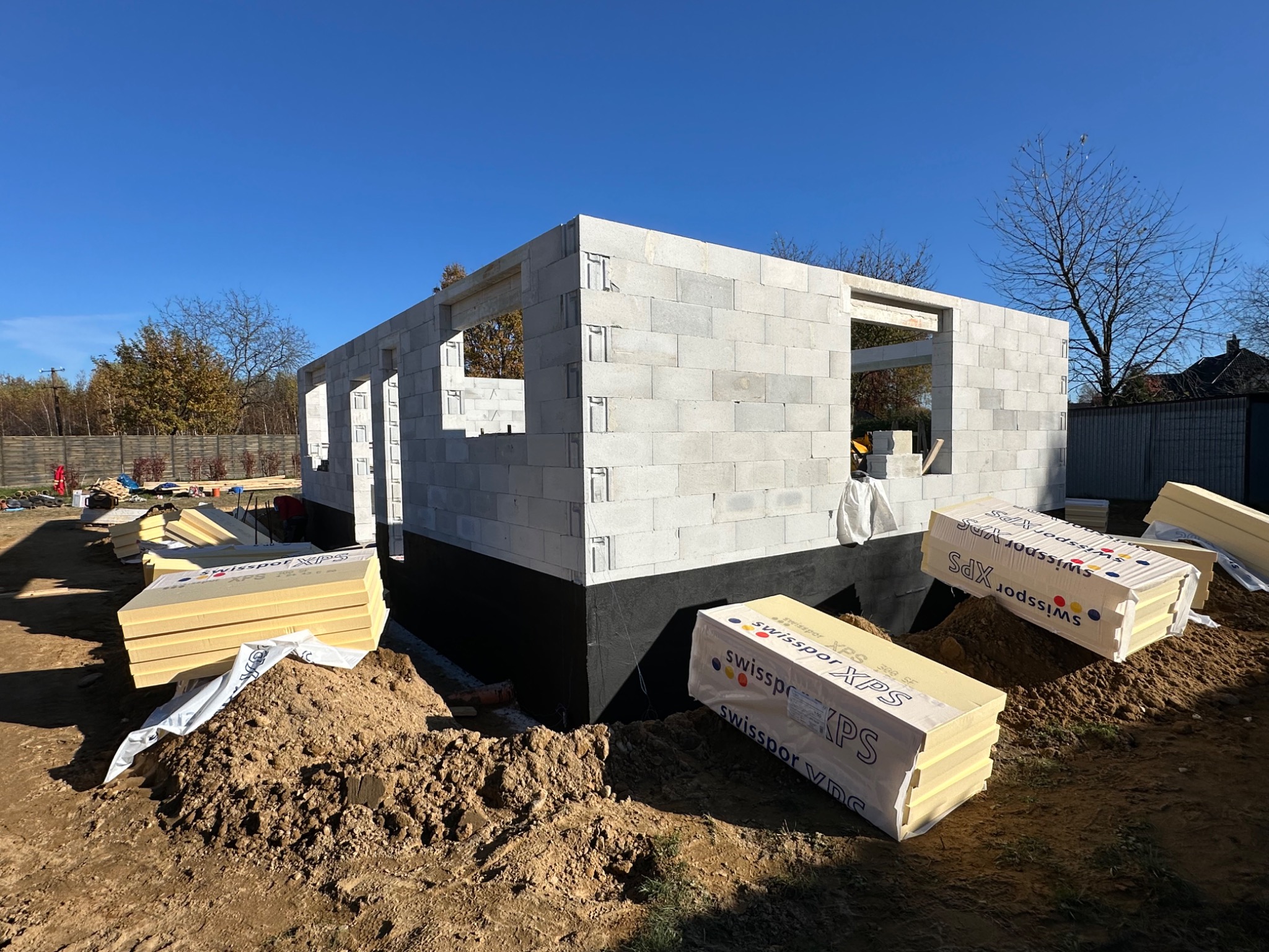 Surowy stan domu z betonu komórkowego, ocieplonego fundamentu i przygotowanymi paczkami styropianu XPS na placu budowy w słoneczny dzień.