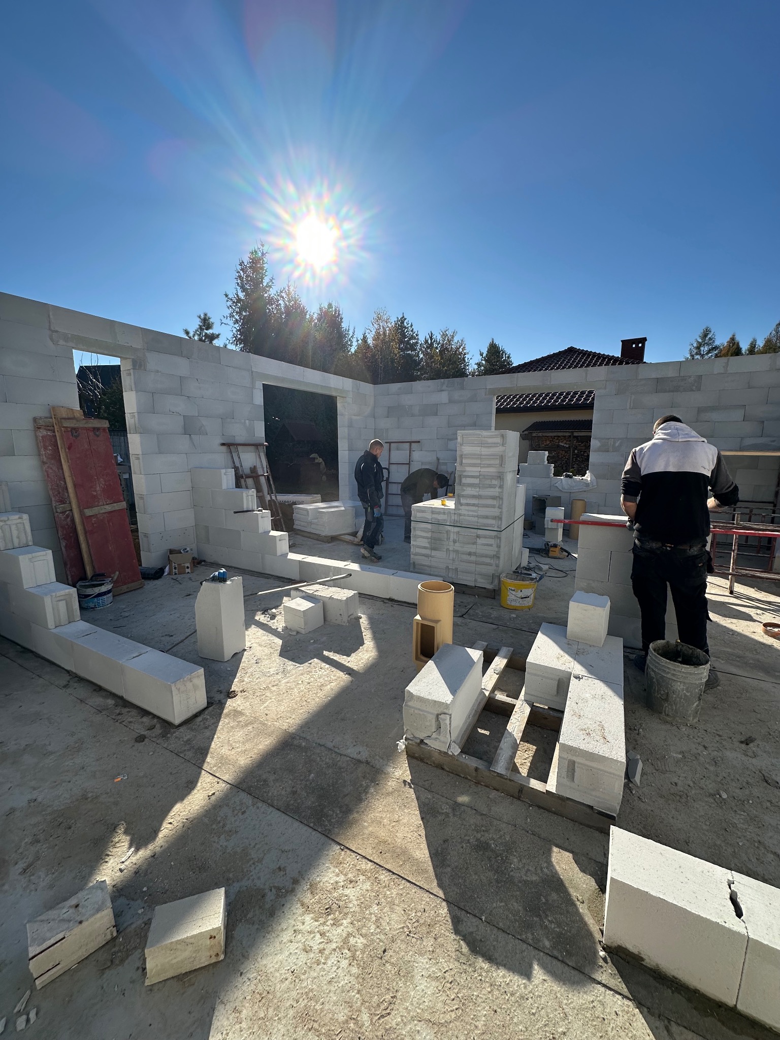 Budowa domu z betonu komórkowego w słoneczny dzień. Widoczne ściany w trakcie wznoszenia, materiały budowlane i dwóch pracowników w tle.