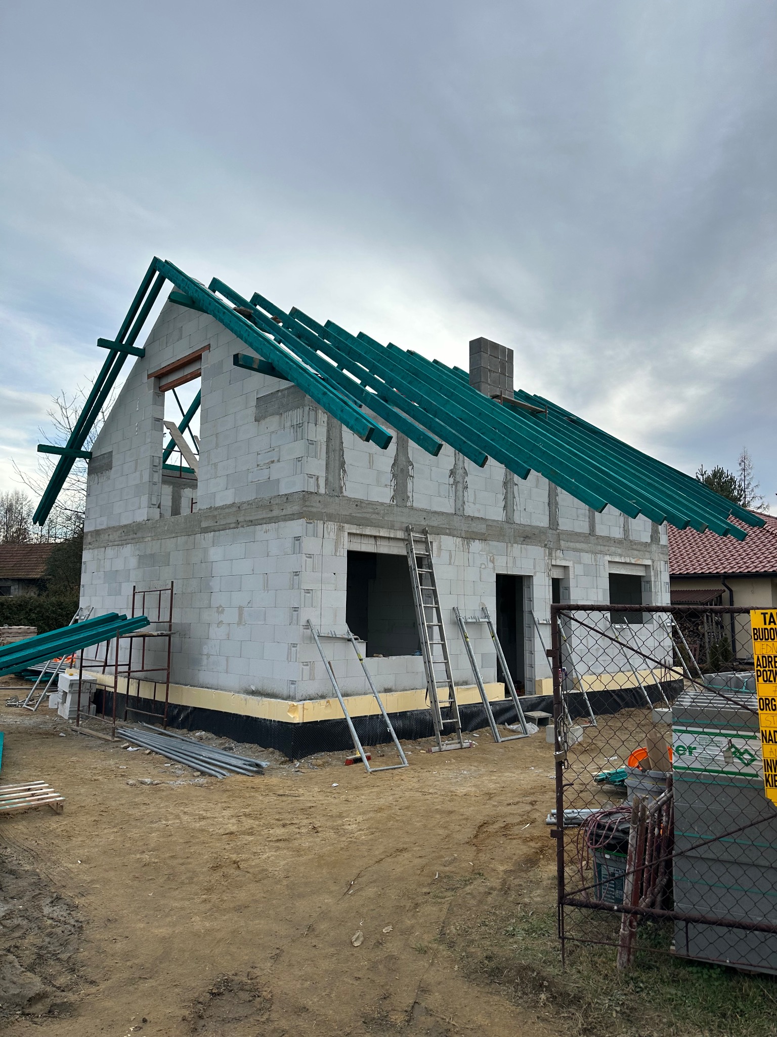 Surowy stan domu z bloczków, z zieloną konstrukcją dachu w trakcie montażu. Widoczne rusztowania, drabiny i materiały budowlane. Dom w stanie deweloperskim.