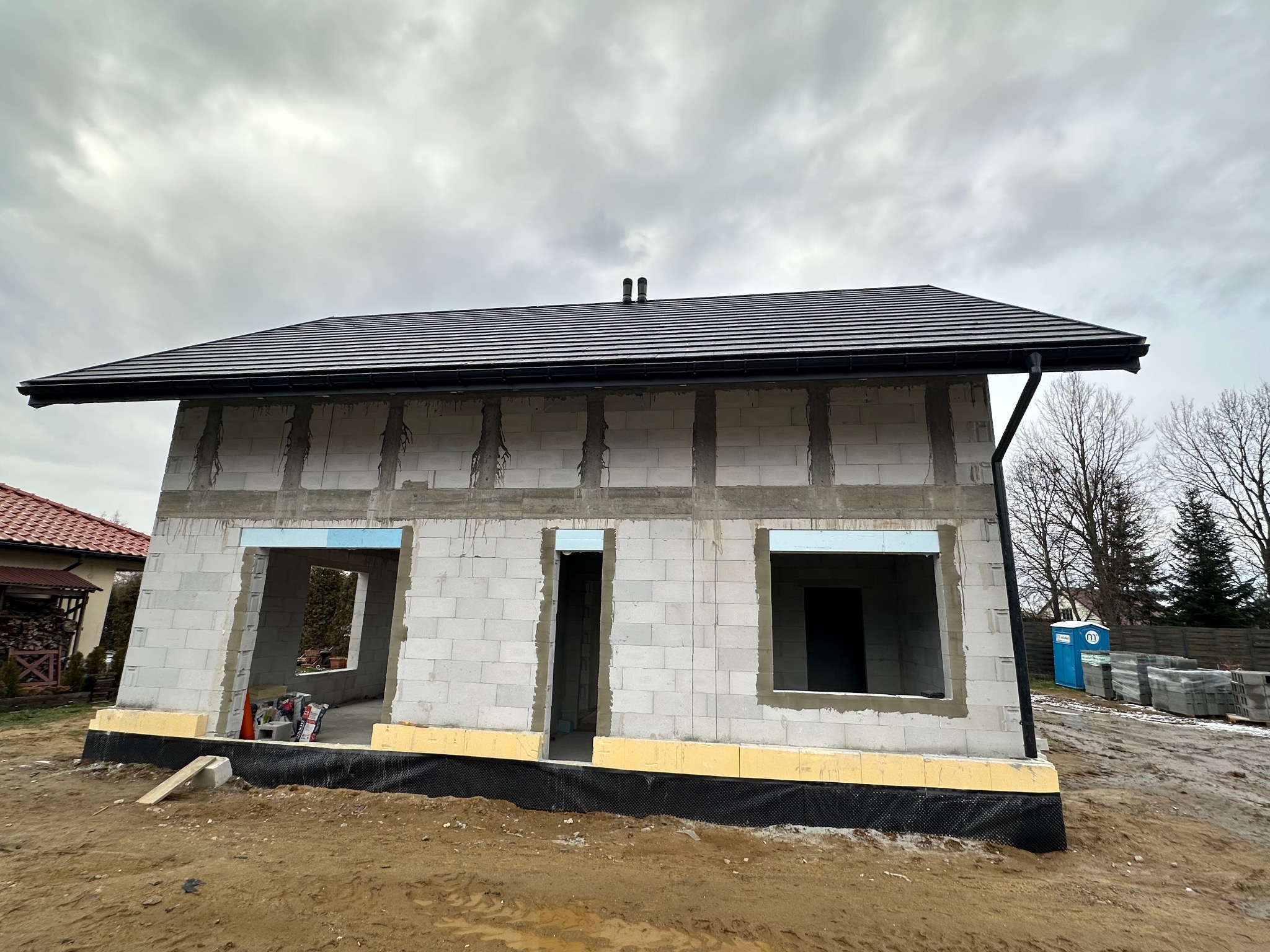 Surowy stan domu z betonu komórkowego z czarnym dachem, widoczne otwory okienne i drzwiowe, ocieplenie fundamentów. Na zewnątrz ziemia i materiały budowlane.