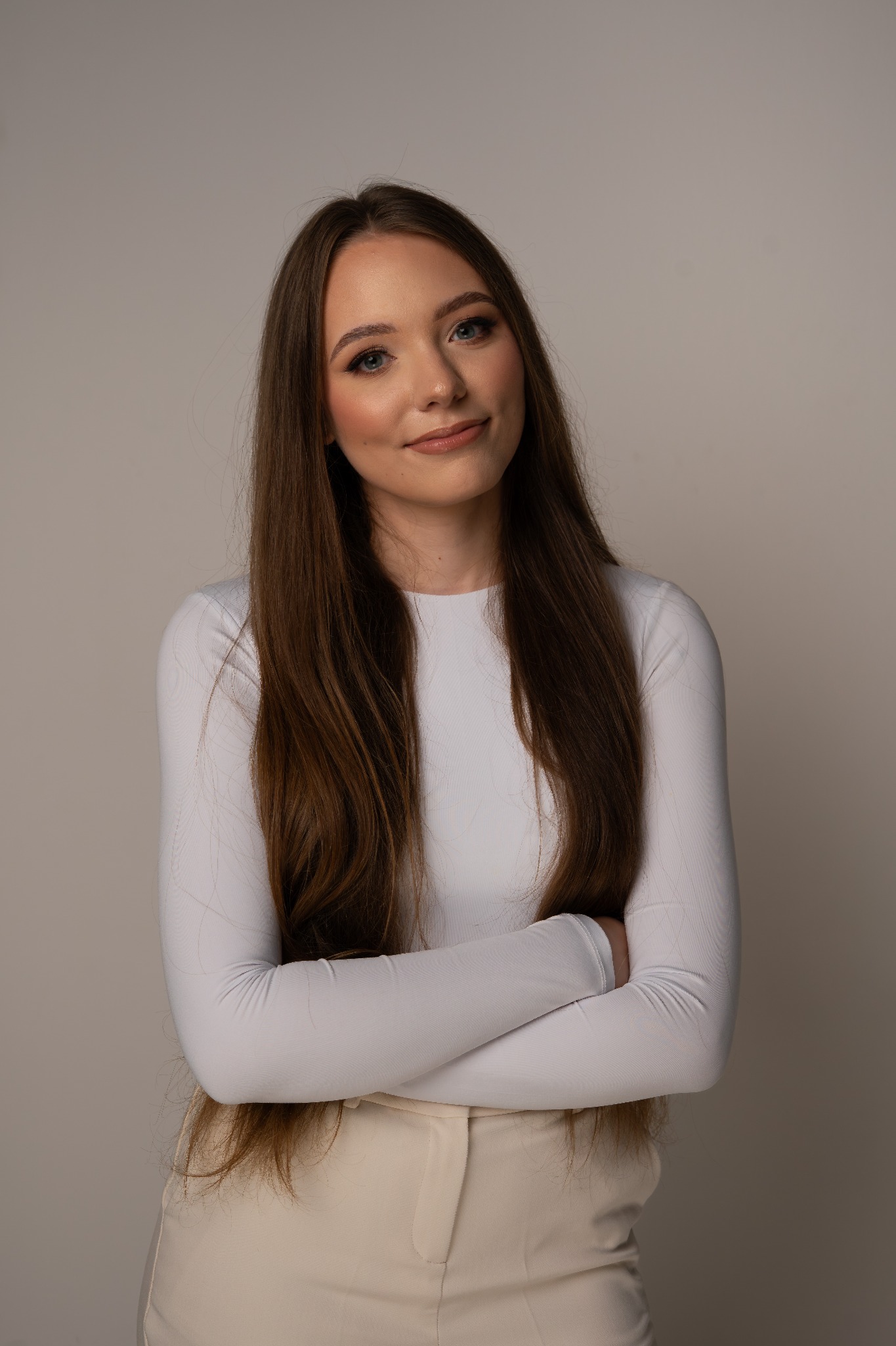 Portret młodej kobiety z długimi brązowymi włosami, ubranej w białą bluzkę i beżowe spodnie, z założonymi rękami na neutralnym tle. Uśmiech na twarzy, delikatny makijaż.