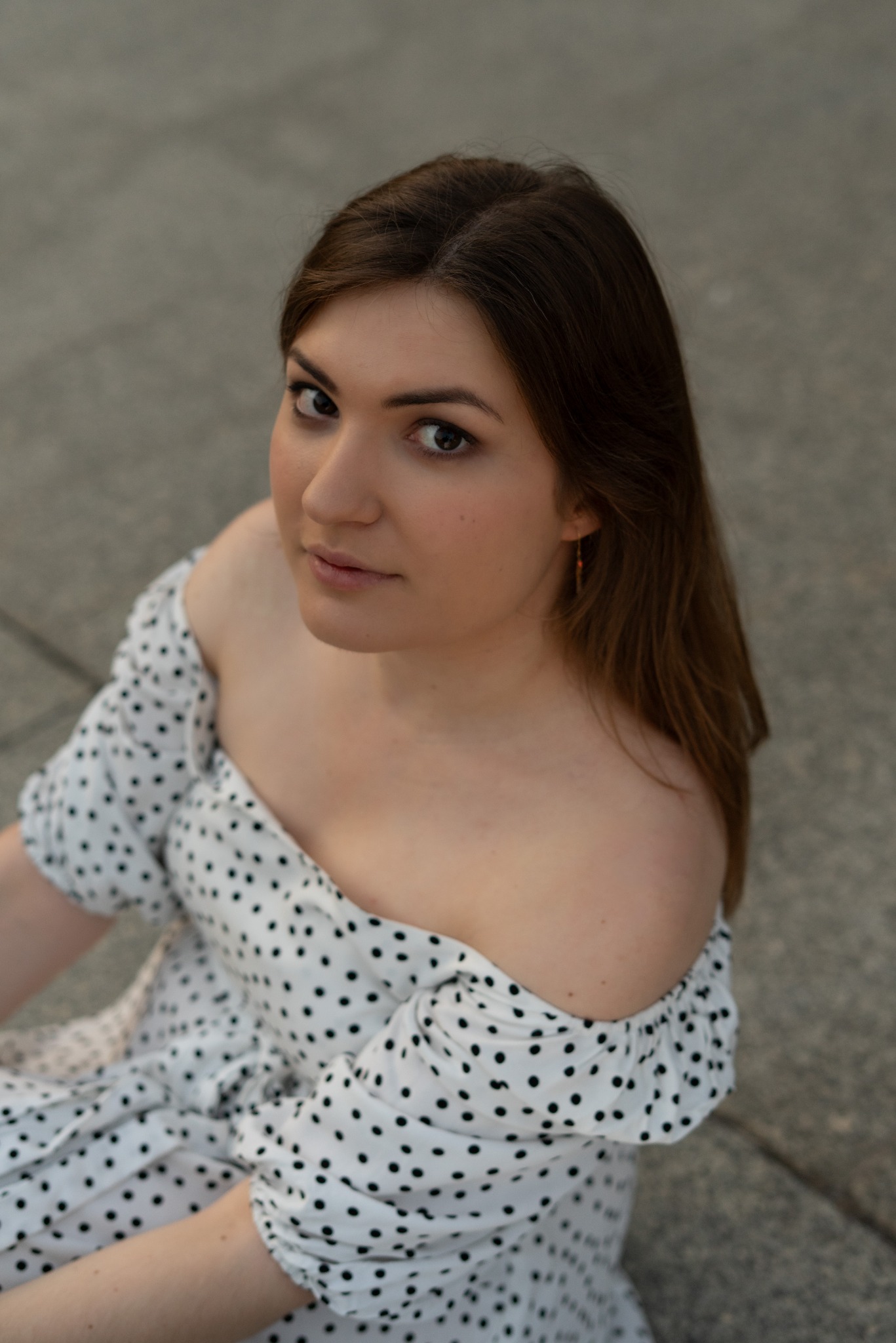 Portret kobiety w sukience w grochy, spoglądającej w górę, z delikatnym uśmiechem i widocznym kolczykiem, na tle szarej powierzchni.