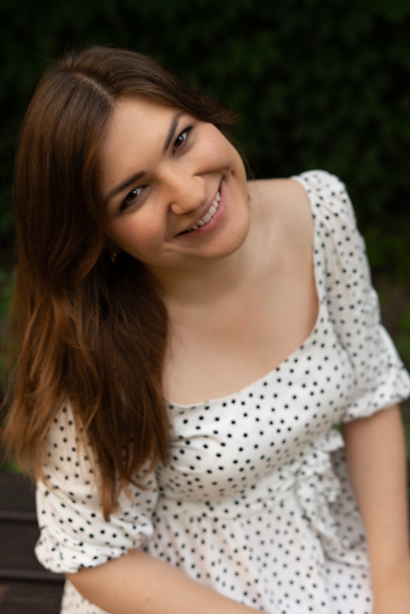 Uśmiechnięta brunetka w sukience w grochy, uchwycona z góry w plenerze w Poznaniu. Naturalne światło, delikatny uśmiech, swobodna poza. Portret.