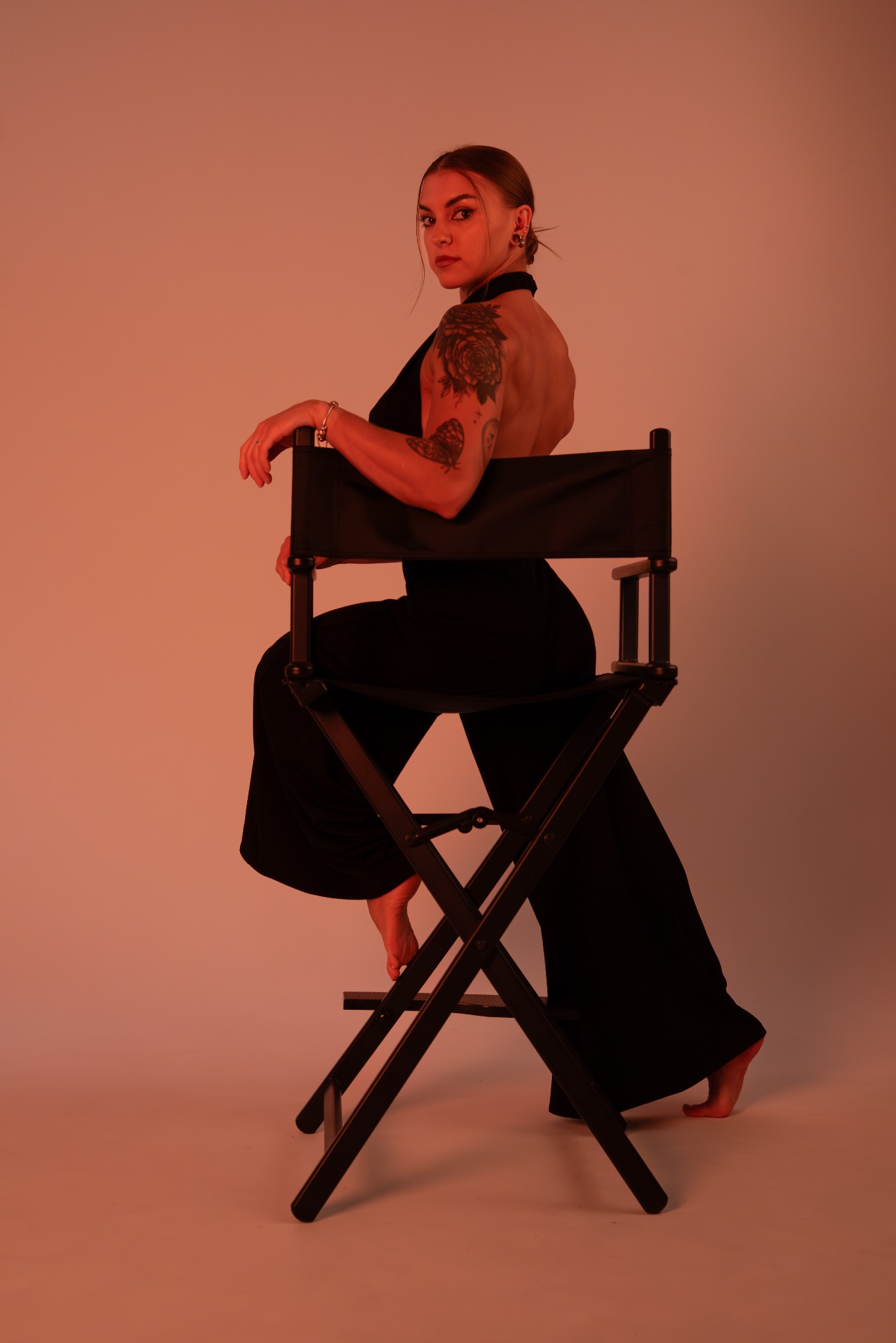 Kobieta w czarnym kombinezonie siedzi na reżyserskim krześle, odwrócona plecami, z tatuażami na ramieniu, w ciepłym oświetleniu studyjnym.