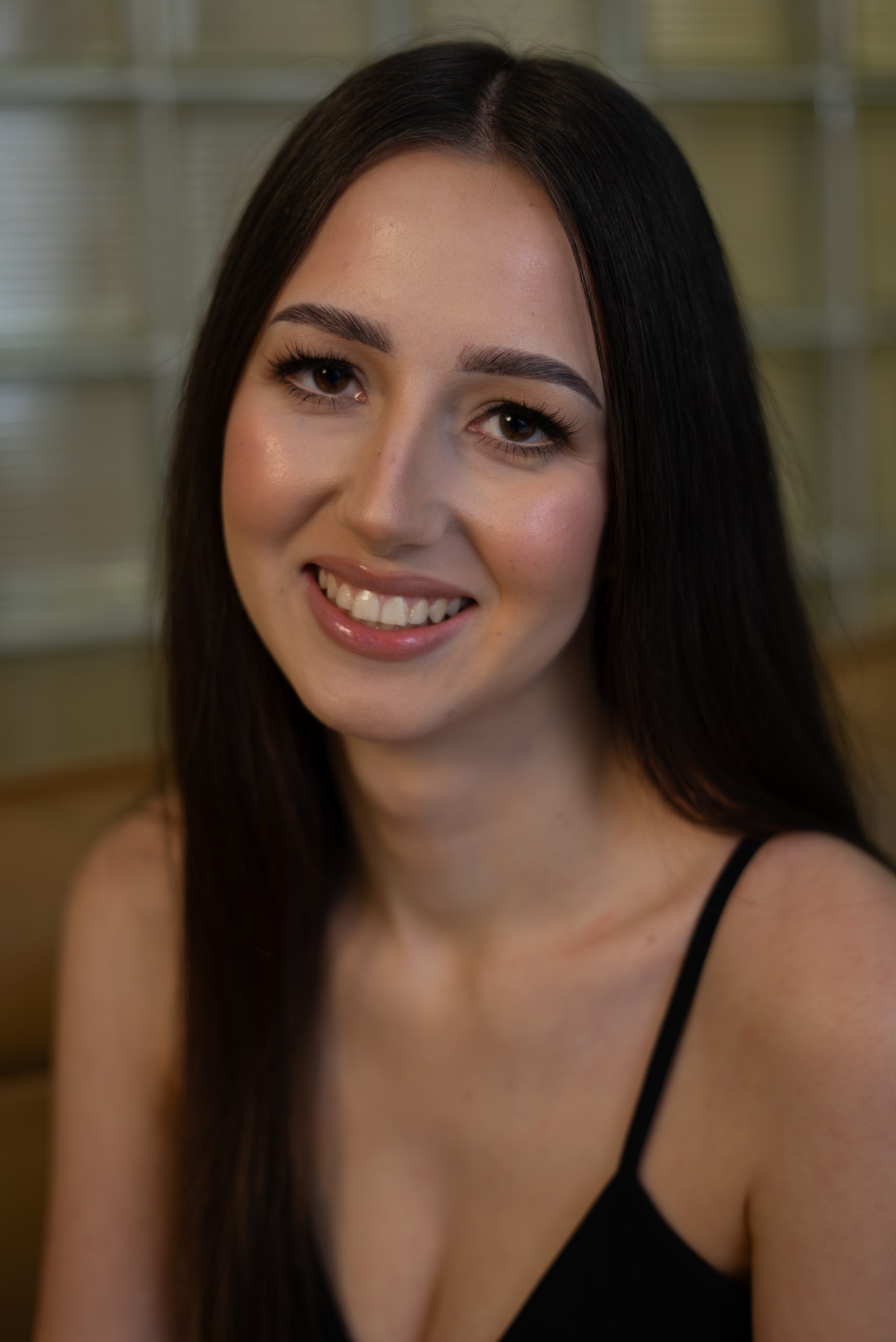 Portret uśmiechniętej, młodej kobiety z długimi, ciemnymi włosami i delikatnym makijażem, na rozmytym tle w ciepłych tonacjach.