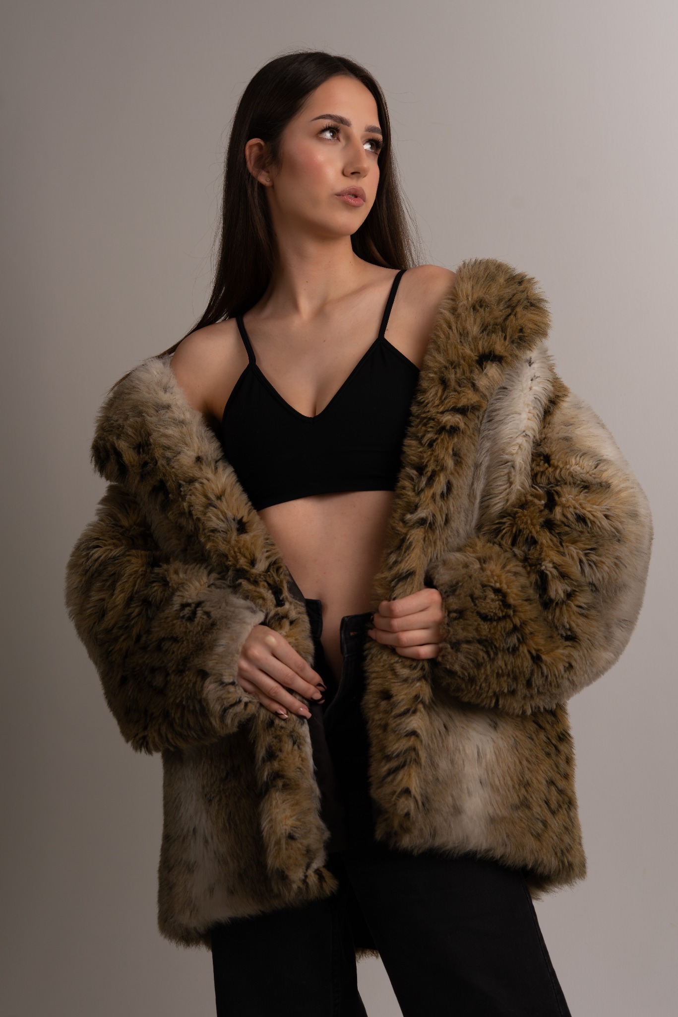 Kobieta w czarnym topie i futrzanym płaszczu, z delikatnym makijażem, pozuje na jasnym tle, spoglądając w bok. Ujęcie studyjne, podkreślające urodę i styl.