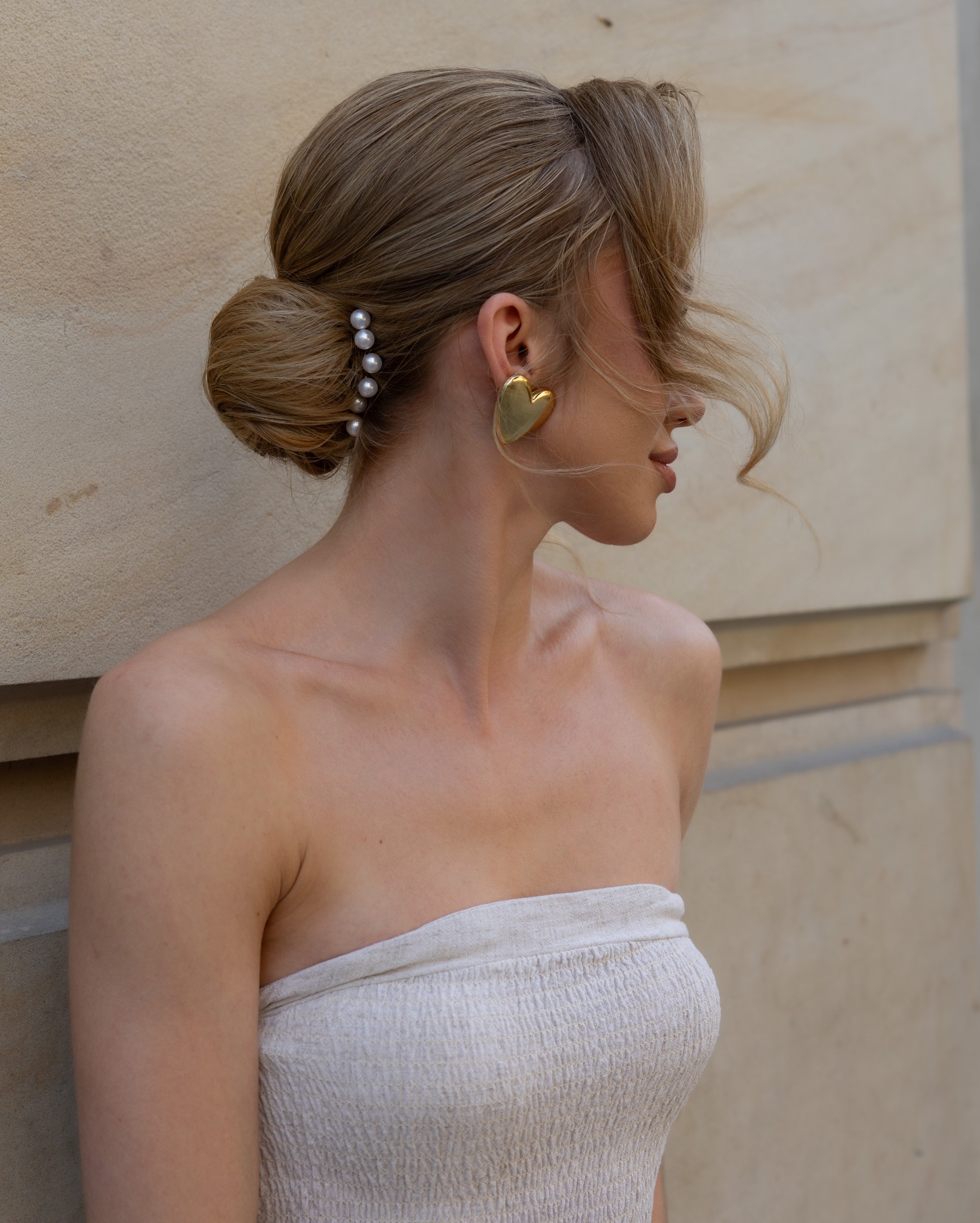 Portret kobiety z upiętymi blond włosami, ozdobionymi perłami, w złotej kolczyku w kształcie serca, na tle beżowej ściany. Profil, delikatne światło.