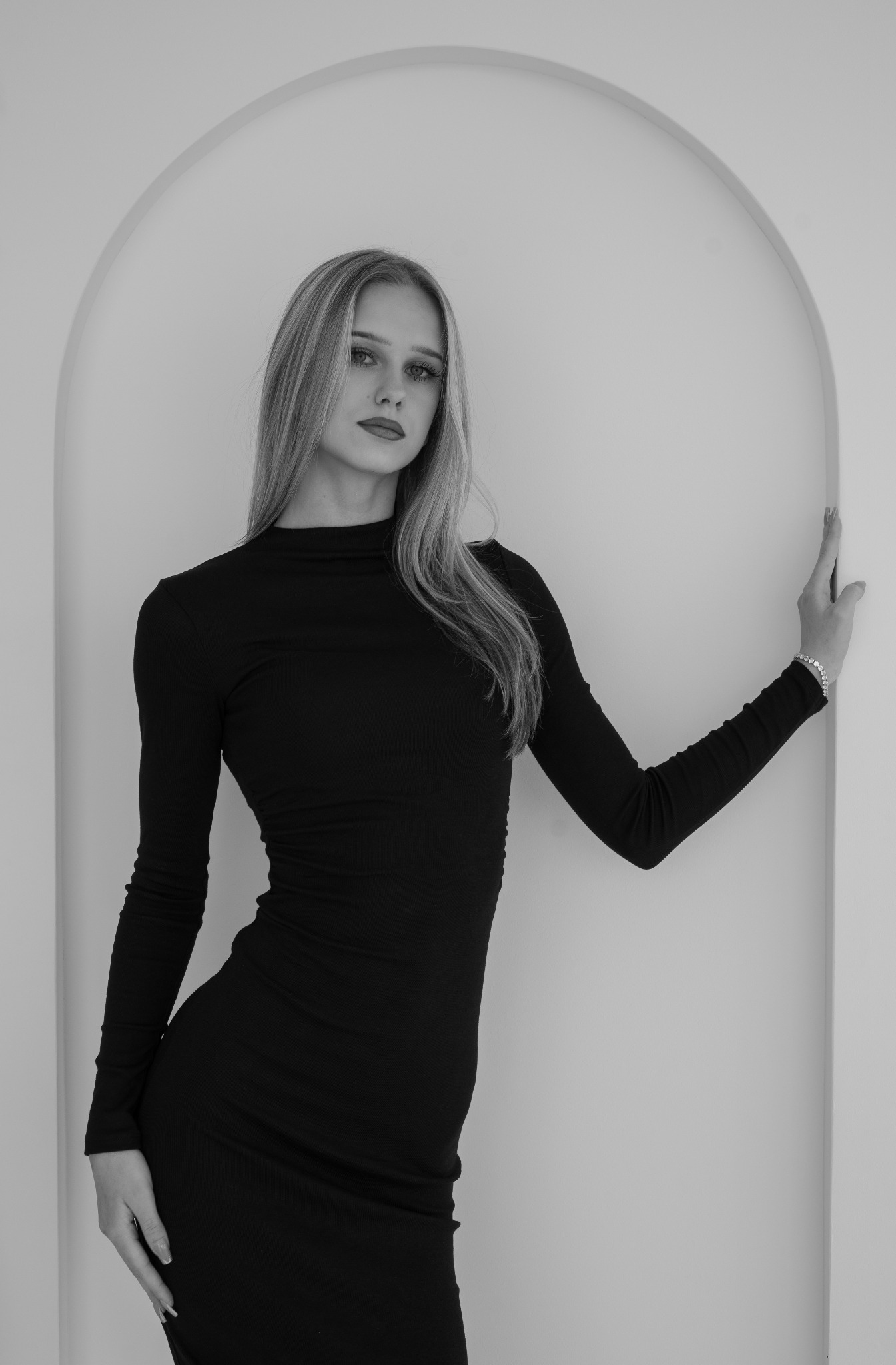 Elegancka, czarno-biała fotografia kobiety w dopasowanej sukience na tle minimalistycznego łuku. Modelka pozuje z ręką opartą o ścianę, eksponując bransoletkę.