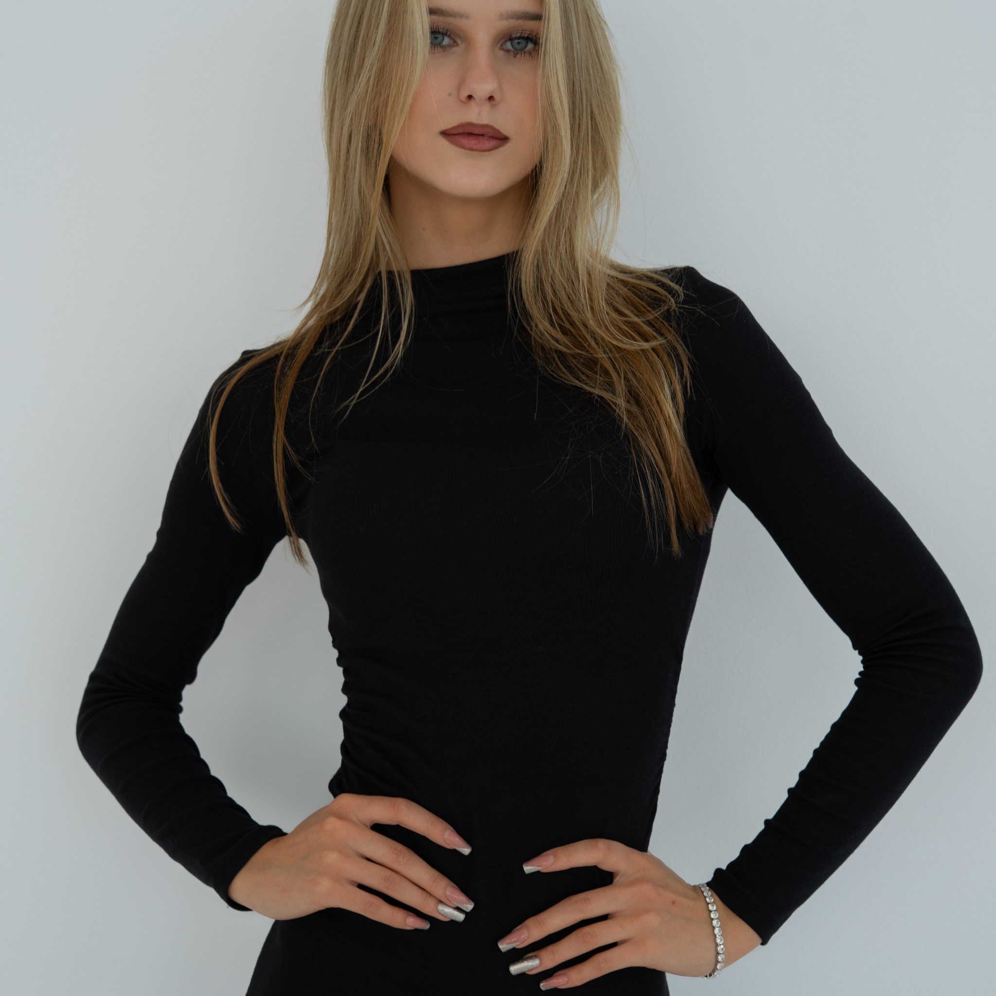 Portret kobiety w czarnej sukience na jasnym tle. Modelka pozuje z rękami na biodrach, prezentując srebrną bransoletkę i manicure. Uwagę przyciągają jej niebieskie oczy.