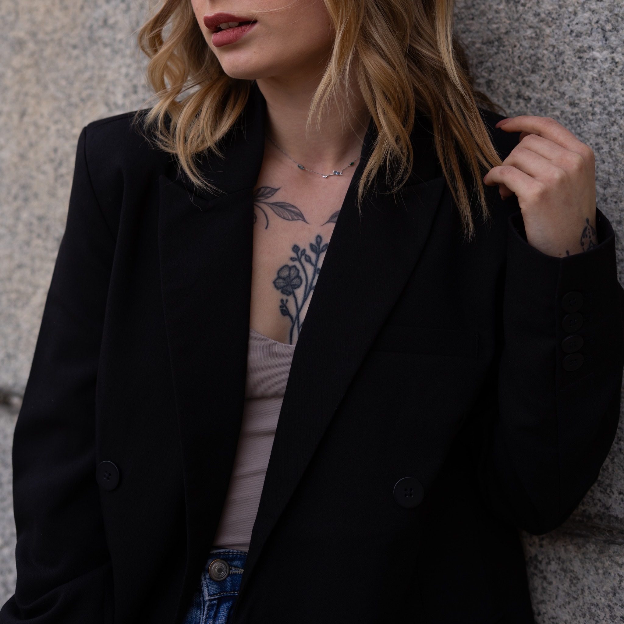 Portret kobiety w czarnej marynarce, z tatuażem na dekolcie, opierającej się o szarą ścianę. Widoczne detale: srebrny naszyjnik i delikatny makijaż.