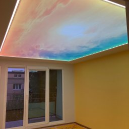 Paweł Czekaj - Pokój z żółtymi ścianami i sufitem napinanym z motywem nieba, oświetlonym listwami LED. Na podłodze folia ochronna. Widok na balkon przez okno.