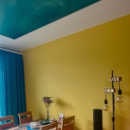 Paweł Czekaj - Żółta ściana i turkusowy sufit z efektem chmur w pokoju w Łodzi. Widoczny stół z dekoracjami i lampa podłogowa. Nietypowe połączenie kolorów.