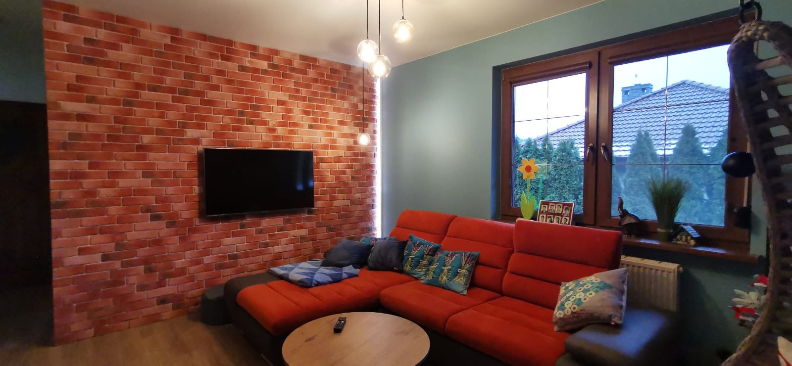 Salon z ceglaną ścianą, na której wisi telewizor. Czerwona sofa z poduszkami, drewniany stolik kawowy i okno z widokiem na zewnątrz. Nowoczesne lampy wiszące.