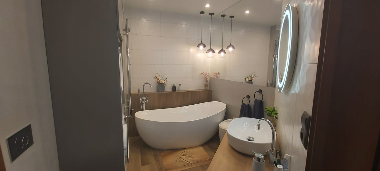 Nowoczesna łazienka z wolnostojącą wanną, umywalką nablatową i okrągłym lustrem z podświetleniem LED. Nad wanną wiszą szklane lampy.