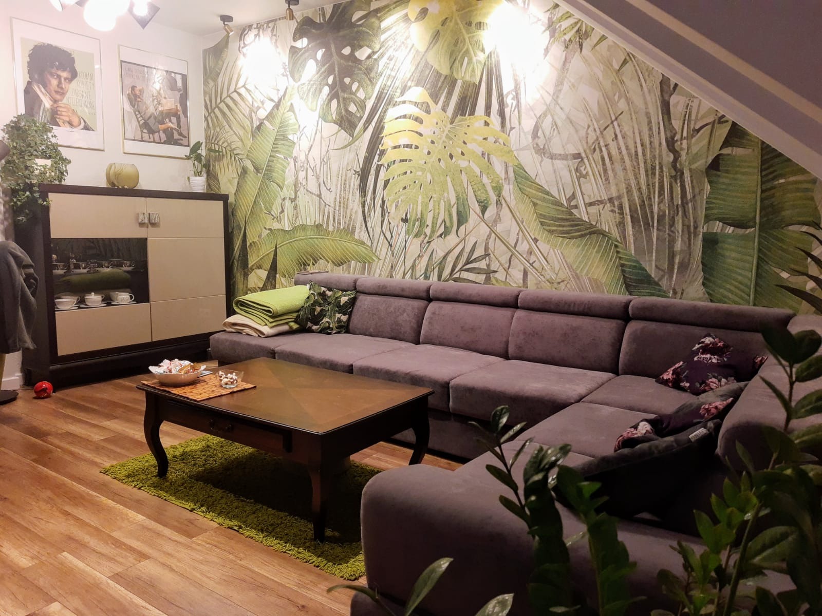 Salon z szarą sofą narożną, drewnianym stolikiem kawowym i tapetą w motywy roślinne. Widoczne elementy dekoracyjne i oświetlenie.