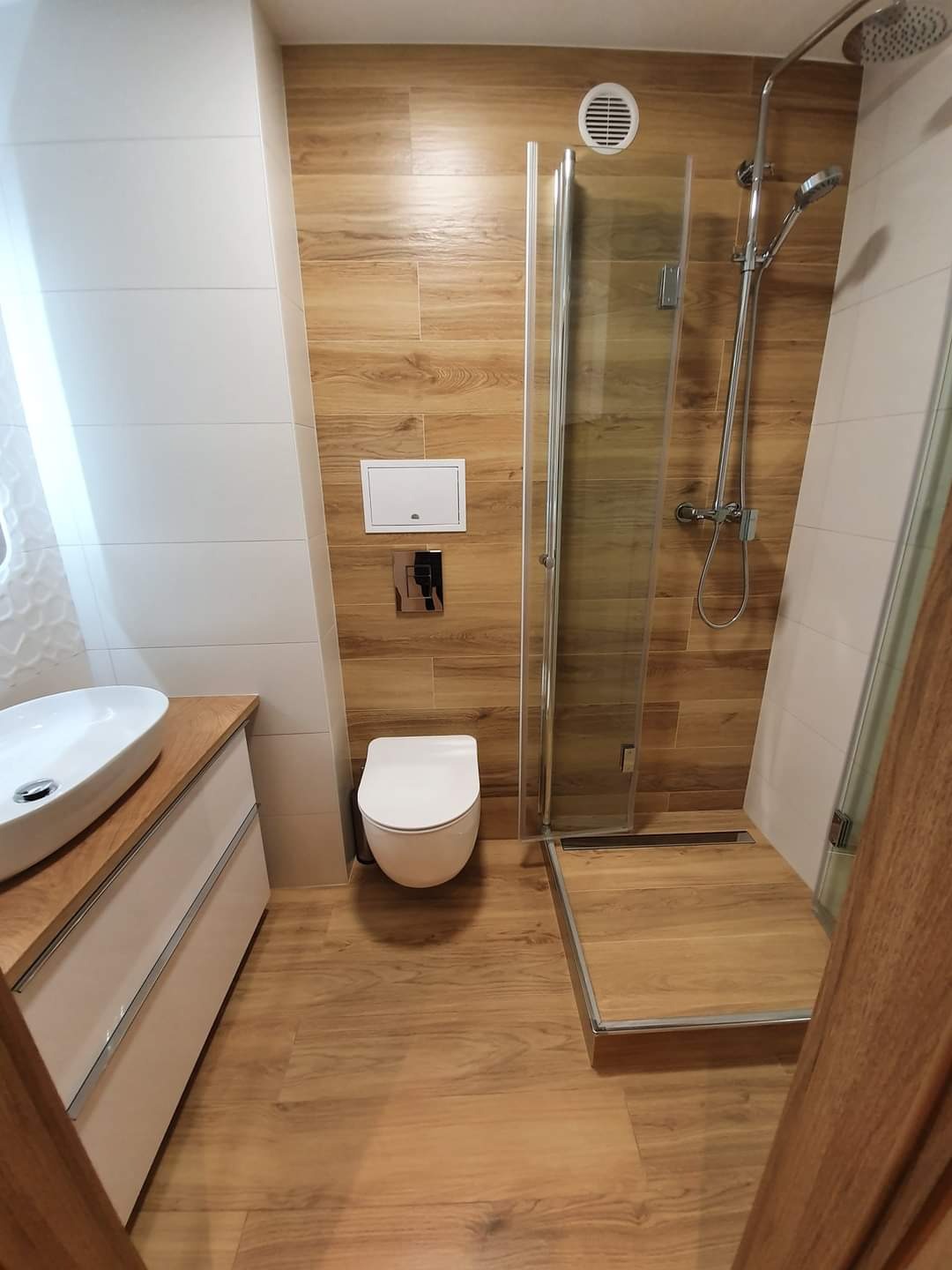 Nowoczesna łazienka z kabiną prysznicową typu walk-in, drewnopodobnymi płytkami na ścianach i podłodze, białą armaturą i szafką pod umywalką z drewnianym blatem.