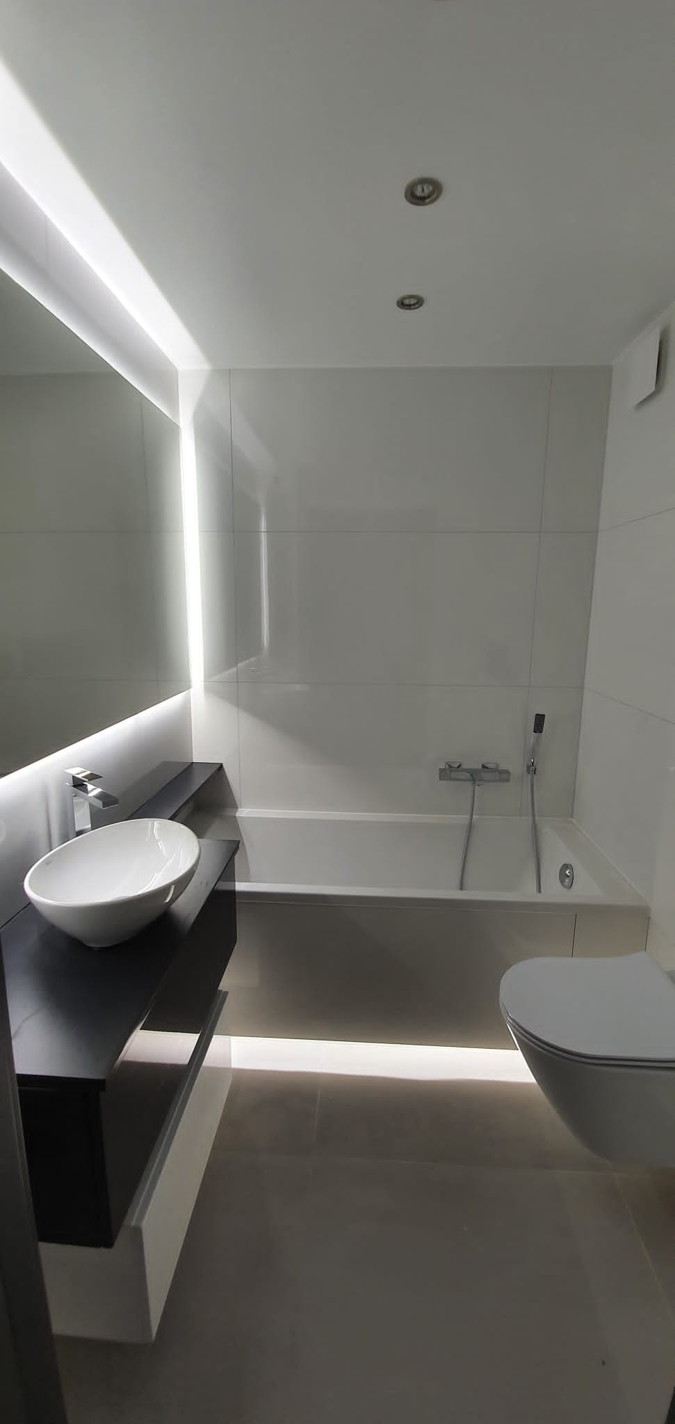 Nowoczesna łazienka z białymi płytkami, podświetlanym lustrem i umywalką nablatową. Minimalistyczny design, oświetlenie punktowe i podświetlenie LED tworzą elegancki klimat.