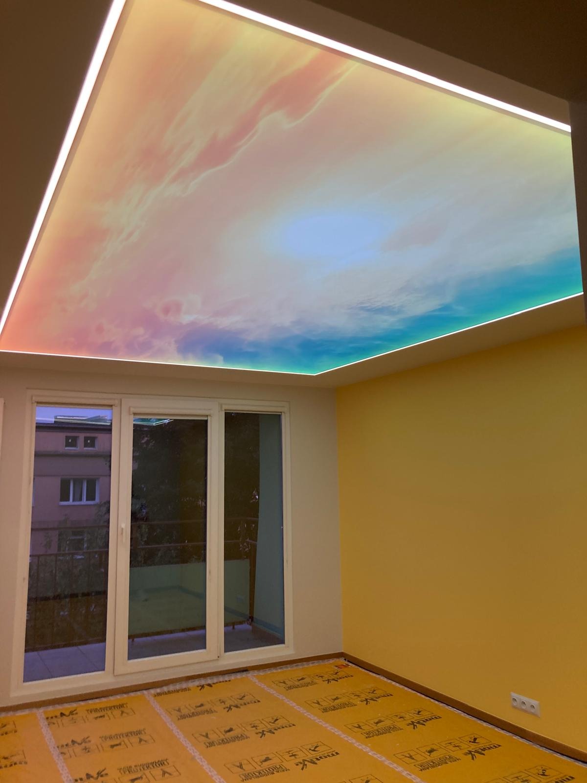 Pokój z żółtymi ścianami i sufitem napinanym z motywem nieba, oświetlonym listwami LED. Na podłodze folia ochronna. Widok na balkon przez okno.
