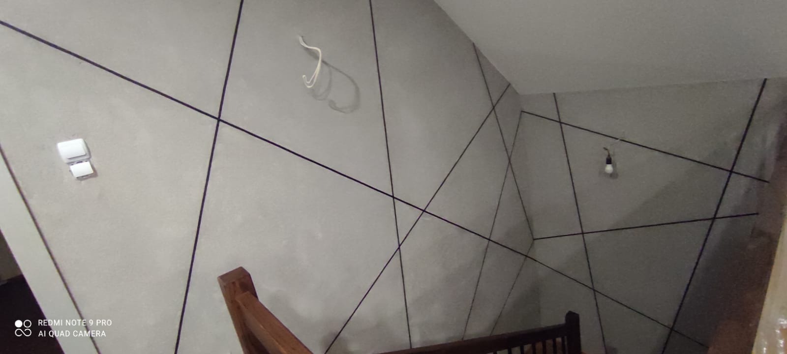 Ściana w szarości z geometrycznym wzorem z czarnych linii, widoczny włącznik światła i fragment schodów. Minimalistyczny design wnętrza.