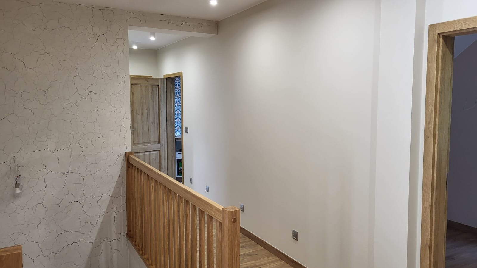 Korytarz po remoncie: ściana z efektem spękań, drewniana balustrada i drzwi. Widoczne oświetlenie sufitowe i drewniana podłoga. Neutralna kolorystyka.