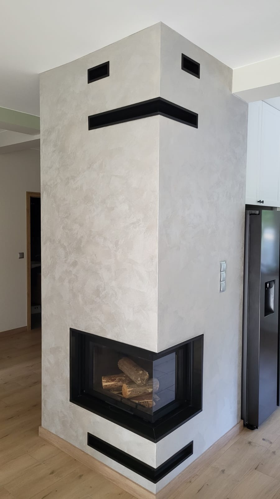 Nowoczesny kominek narożny z czarną obudową i dekoracyjną, szarą strukturą tynku. Widoczne drewno w palenisku, fragment kuchni w tle. Minimalistyczny design.
