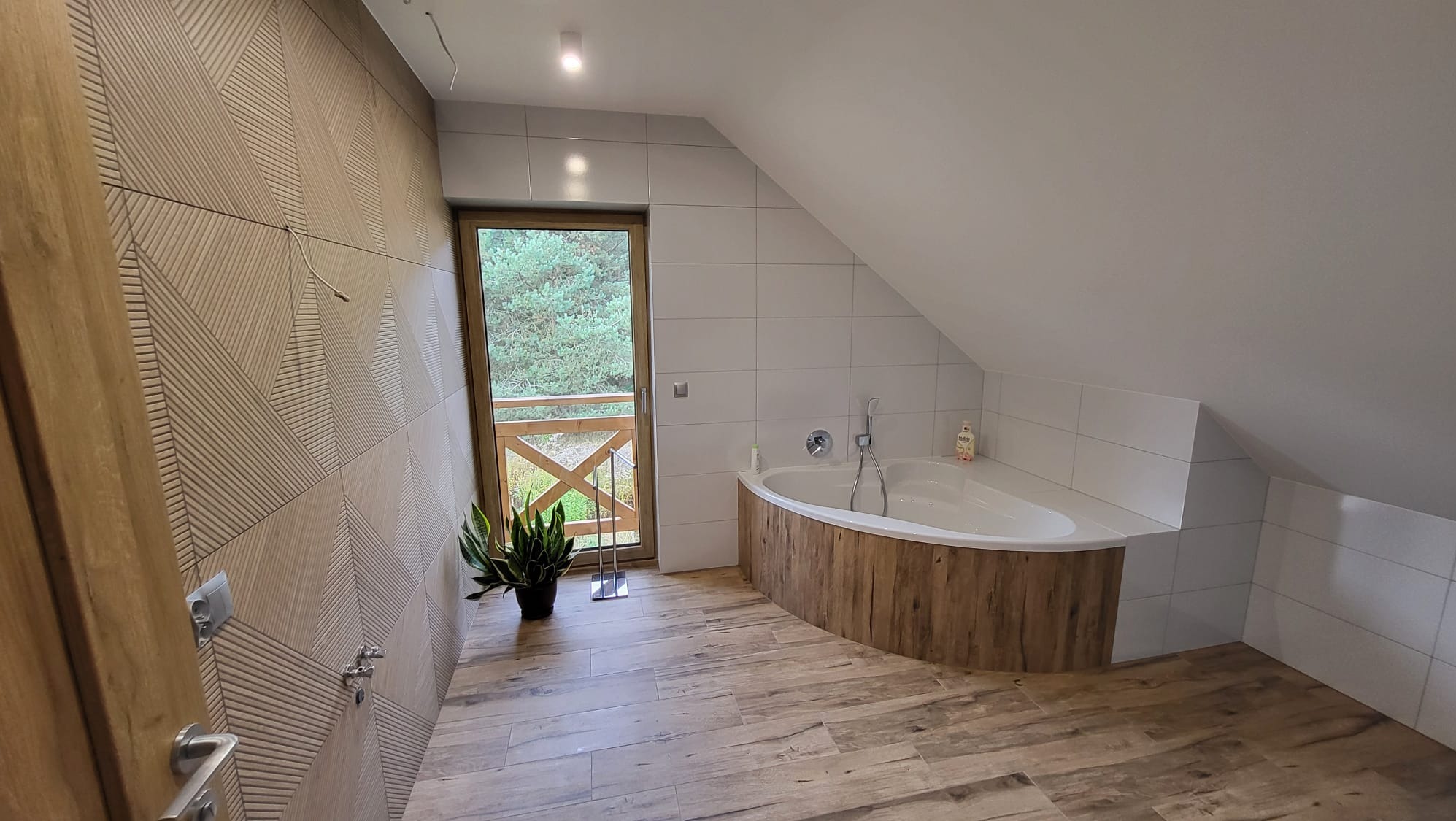 Wykończona łazienka na poddaszu z wanną obudowaną drewnem, jasnymi płytkami i drewnopodobną podłogą. Widok na zieleń przez okno balkonowe.
