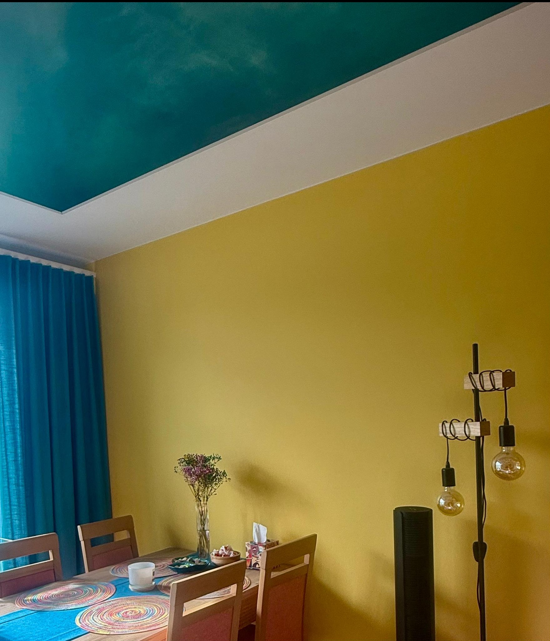 Żółta ściana i turkusowy sufit z efektem chmur w pokoju w Łodzi. Widoczny stół z dekoracjami i lampa podłogowa. Nietypowe połączenie kolorów.