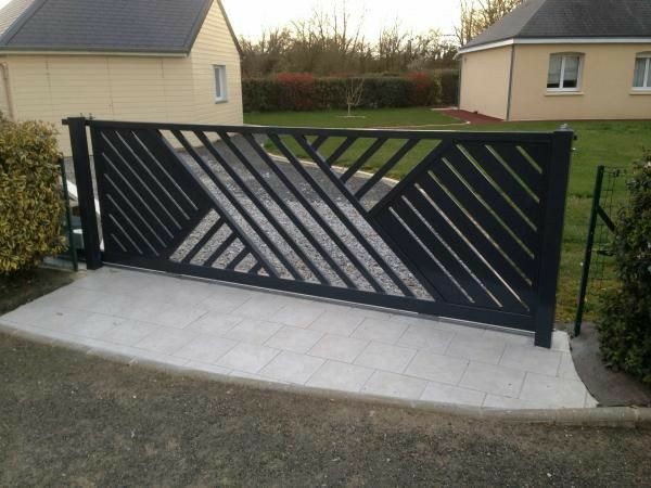 Nowoczesna, przesuwna brama metalowa w kolorze antracytu z geometrycznym wzorem na tle domu i zieleni. Brama zamontowana na jasnej, wyłożonej płytkami nawierzchni.