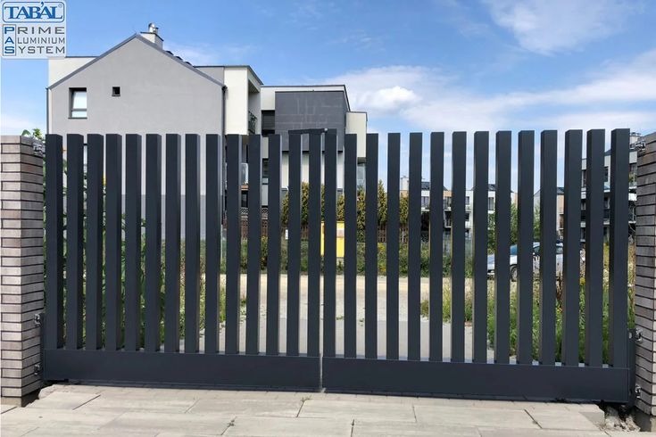 Nowoczesna, przesuwna brama metalowa z pionowymi szczeblami, osadzona między murowanymi słupkami. W tle widoczny budynek mieszkalny i błękitne niebo.