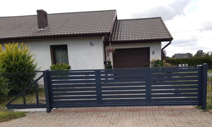 Nowoczesna, przesuwna brama w kolorze antracytowym z poziomymi panelami, na tle domu z brązowym dachem i kostki brukowej. Widoczny fragment ogrodzenia panelowego.