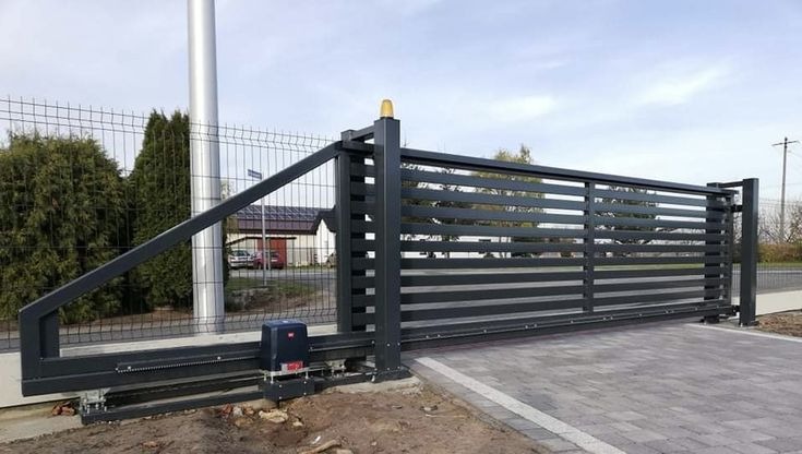 Nowoczesna, przesuwna brama wjazdowa z automatyką, pomalowana na grafitowo, z poziomymi szczeblami, na tle ogrodzenia z siatki i kostki brukowej.