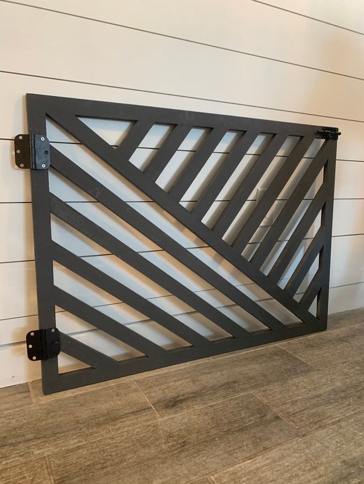 Szara, drewniana bramka zabezpieczająca z ukośnymi szczebelkami, zamontowana na zawiasach, na tle białej ściany z boazerią i podłogi z płytek imitujących drewno.