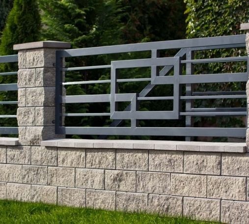 Nowoczesne ogrodzenie metalowe z geometrycznym wzorem, osadzone na murku z kamienia łupanego. Szara kolorystyka, trawnik w tle.