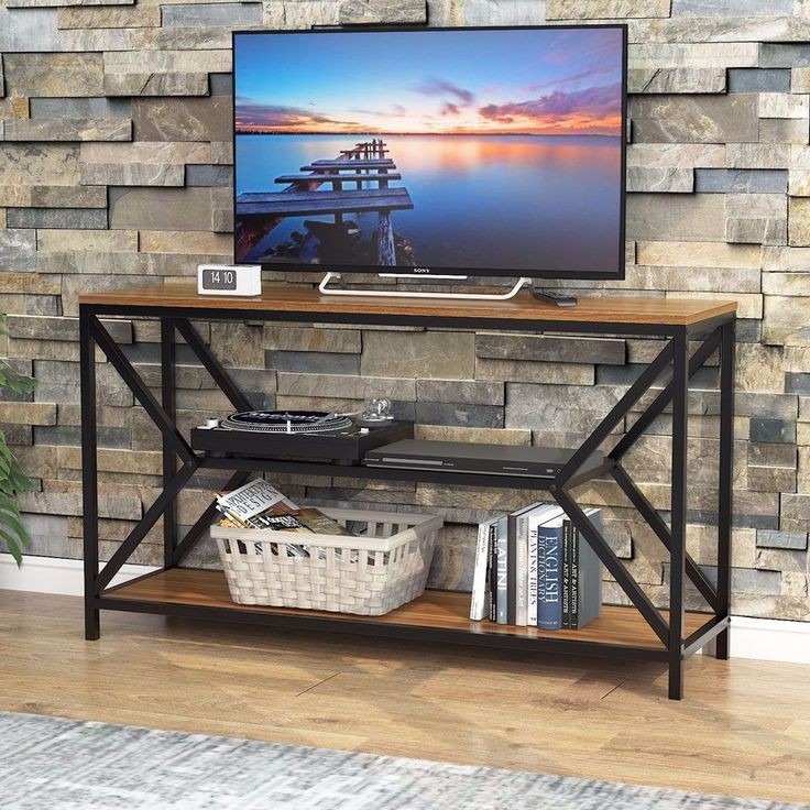 Stolik RTV z metalową ramą i drewnianymi półkami, na tle ściany z kamienia dekoracyjnego. Na blacie telewizor, na półkach książki, płyty i dekoracje.