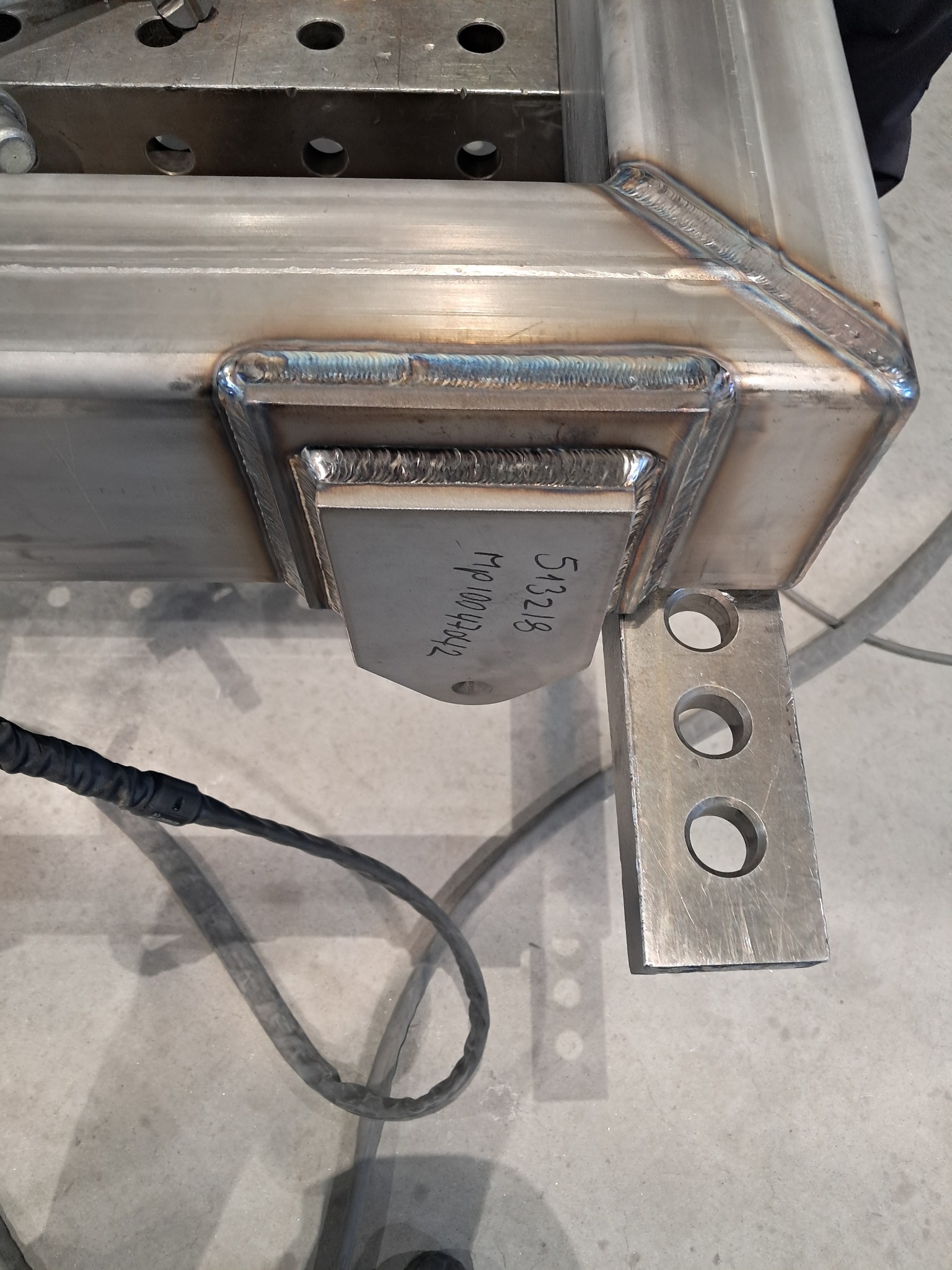 Detal spawu ramy ze stali nierdzewnej z przymocowanym elementem z otworami, widoczne ślady po spawaniu i numer seryjny na elemencie mocującym.