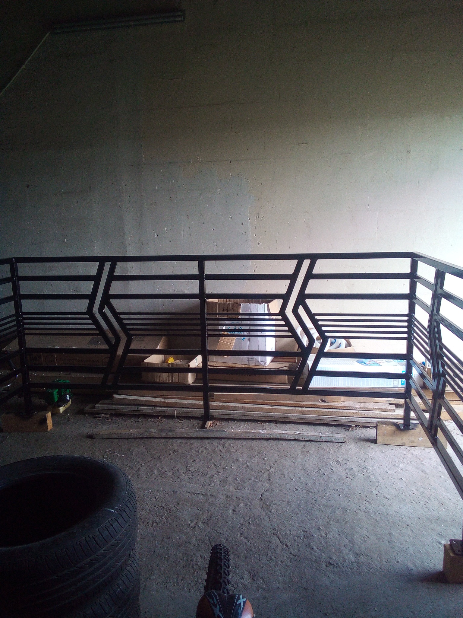 Czarna balustrada metalowa z geometrycznym wzorem, widoczna z dołu, w tle szara ściana i kartony. Elementy opony i roweru w dolnej części kadru.