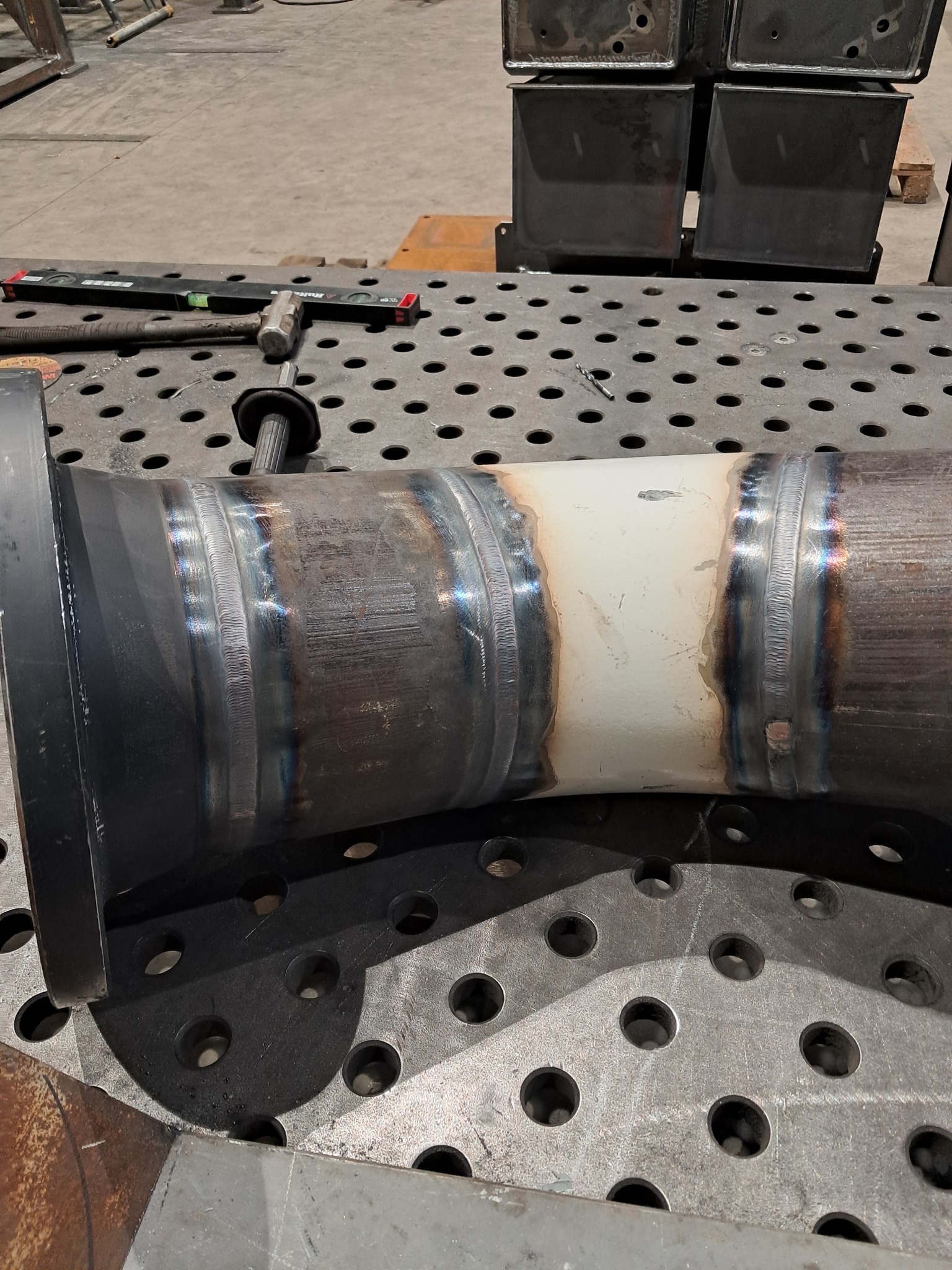 Spoina na rurze z kołnierzem, widoczna na perforowanym stole spawalniczym. Ślady obróbki i narzędzia w tle. Detal spawalniczy w warsztacie.