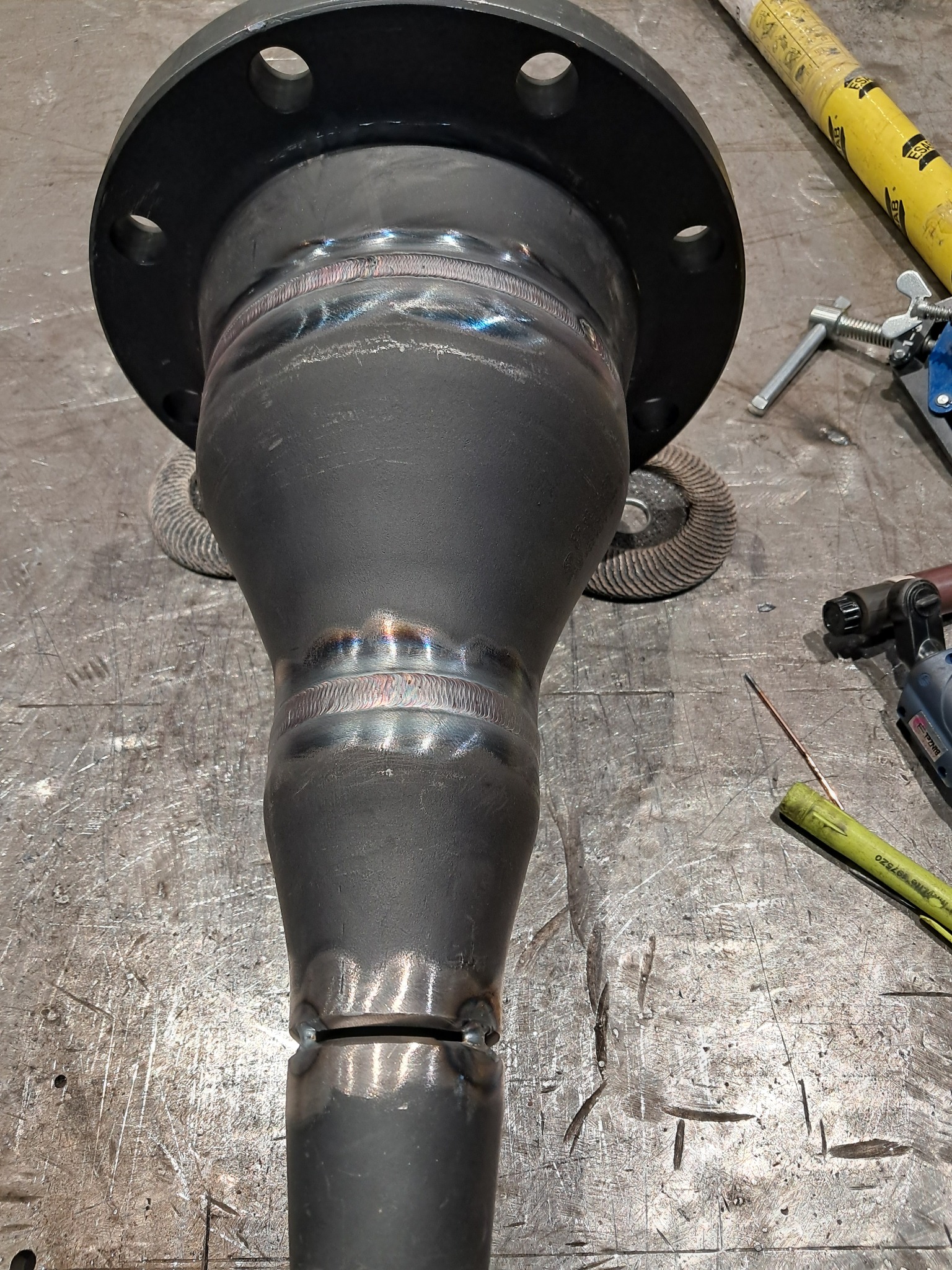 Metalowa rura z kołnierzem po spawaniu, widoczne spoiny i narzędzia na metalowym stole warsztatowym. Detal z bliska, ukazujący precyzję wykonania.