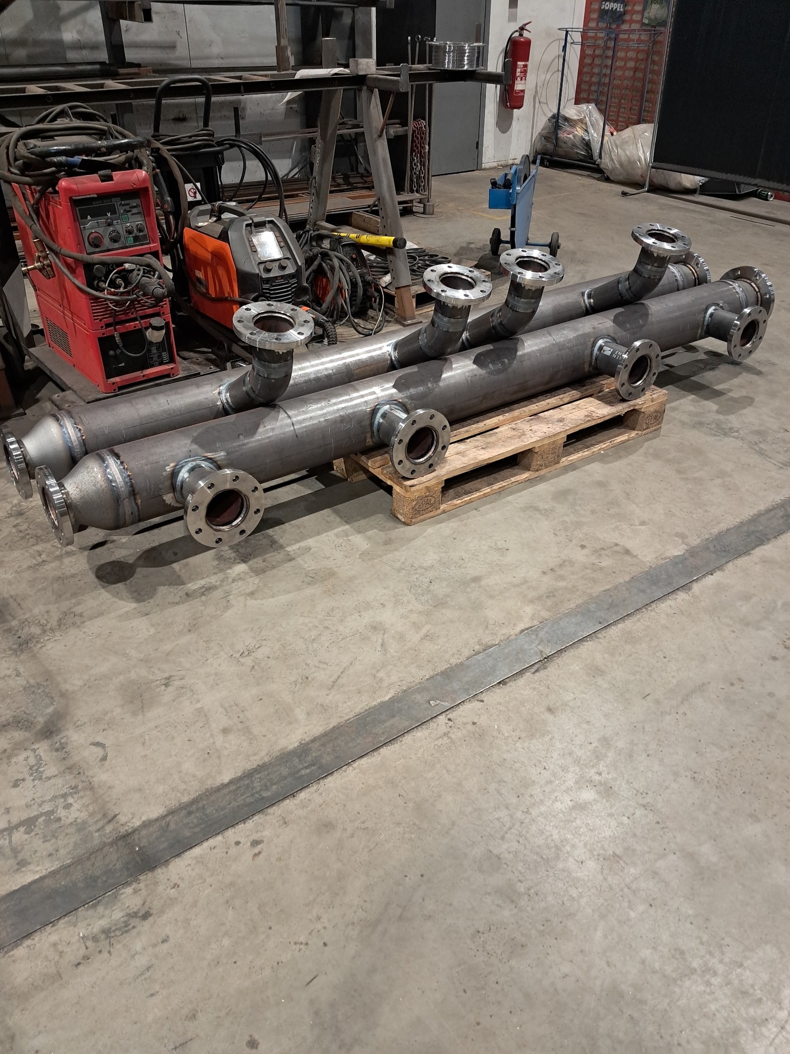 Dwa metalowe kolektory rurowe z przyspawanymi kołnierzami na palecie, obok spawarki w warsztacie. Widoczne ślady spawania i obróbki metalu.