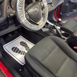 NZK AUTO DETAILING - Olszewnica Stara - Wnętrze czerwonego auta z pokrowcem na kierownicy i papierową matą pod nogami. Widoczne czarne fotele i deska rozdzielcza. Przygotowanie do czyszczenia.