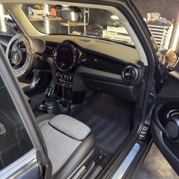 NZK AUTO DETAILING - Olszewnica Stara - Wnętrze czarnego Mini Coopera po detailingu: czyste fotele z szarej tkaniny, dywaniki i deska rozdzielcza. Kierownica zabezpieczona folią. Widoczne narzędzia w tle.