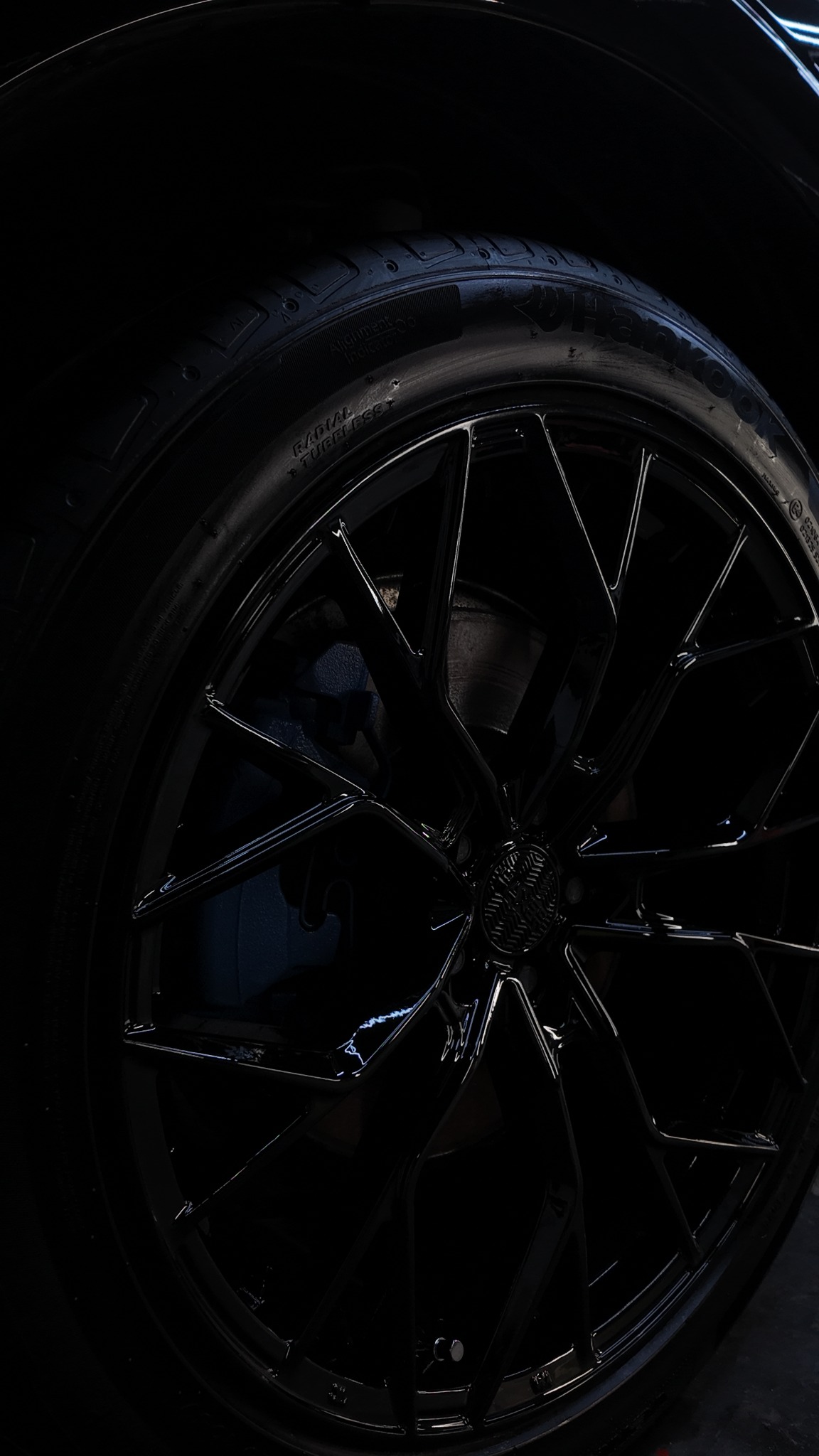 Błyszczące, czarne felgi aluminiowe z oponą na samochodzie, widoczne z bliska pod kątem, z detalami bieżnika i wzoru felgi. Ciemne tło podkreśla połysk.