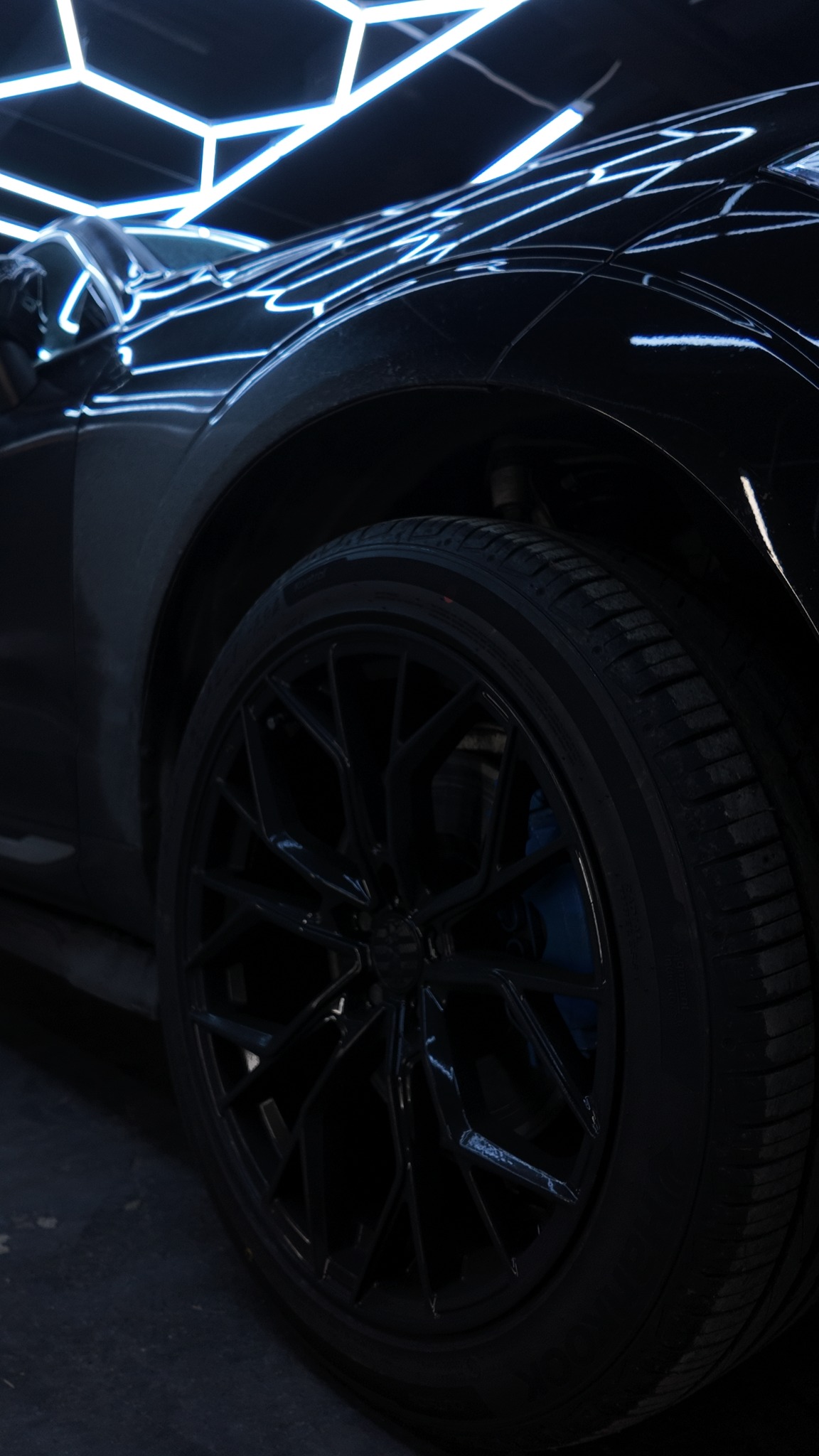 Częściowy widok czarnego samochodu z aluminiowymi felgami i niebieskimi zaciskami hamulcowymi, oświetlonego neonowym światłem w kształcie plastra miodu.