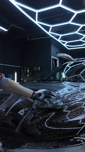 Czyszczenie karoserii czarnego auta gąbką w rękawicy, wnętrze myjni z oświetleniem LED. Piana na lakierze, detale mycia.