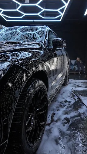 Czarny SUV pokryty pianą w myjni, widok z boku, oświetlenie LED w kształcie plastra miodu, osoba w tle. Detailing felg i karoserii.