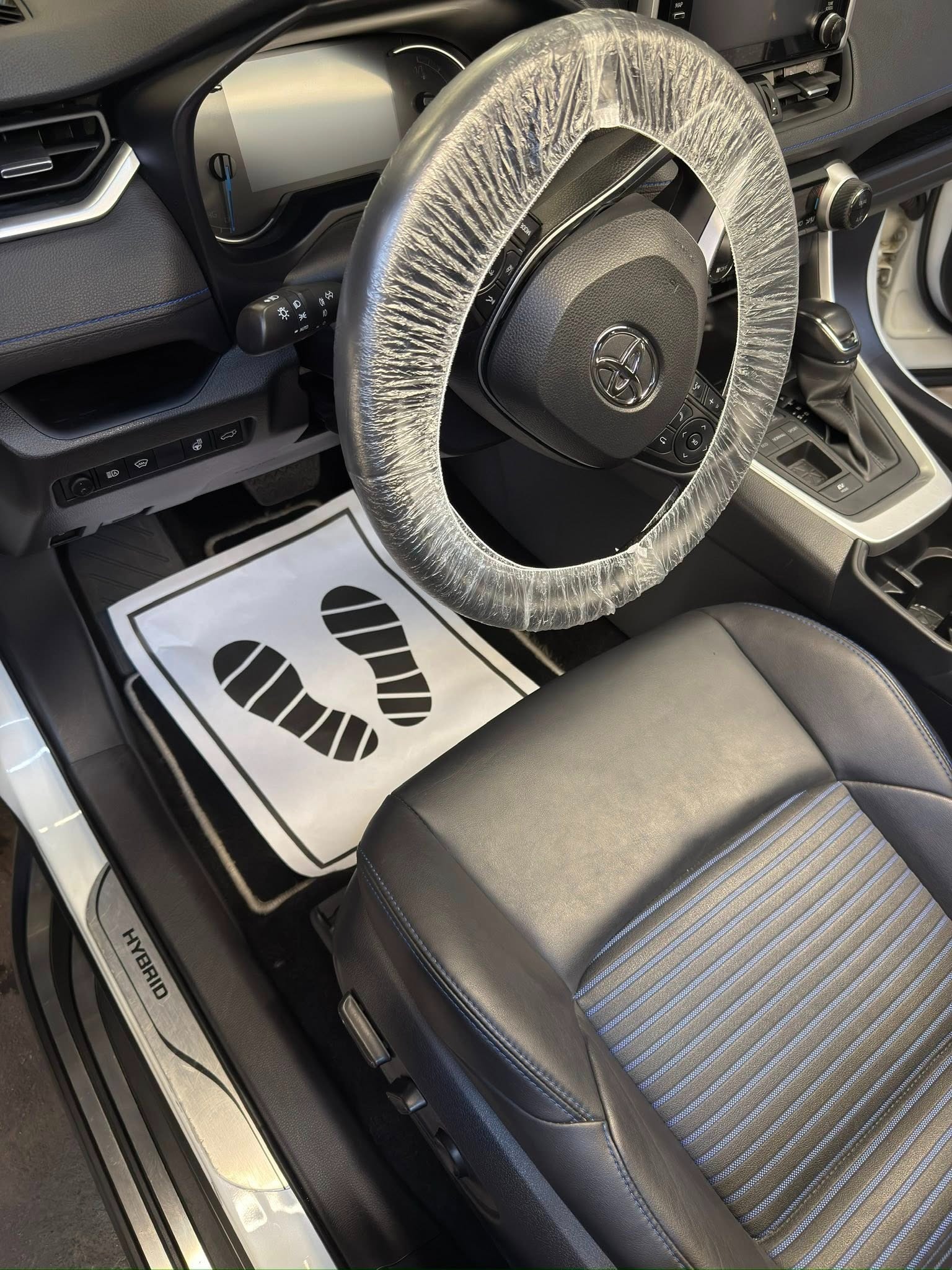 Wnętrze hybrydowego auta z założonym pokrowcem na kierownicy i papierową matą pod nogami. Widoczne czarne skórzane siedzenie z niebieskimi paskami.