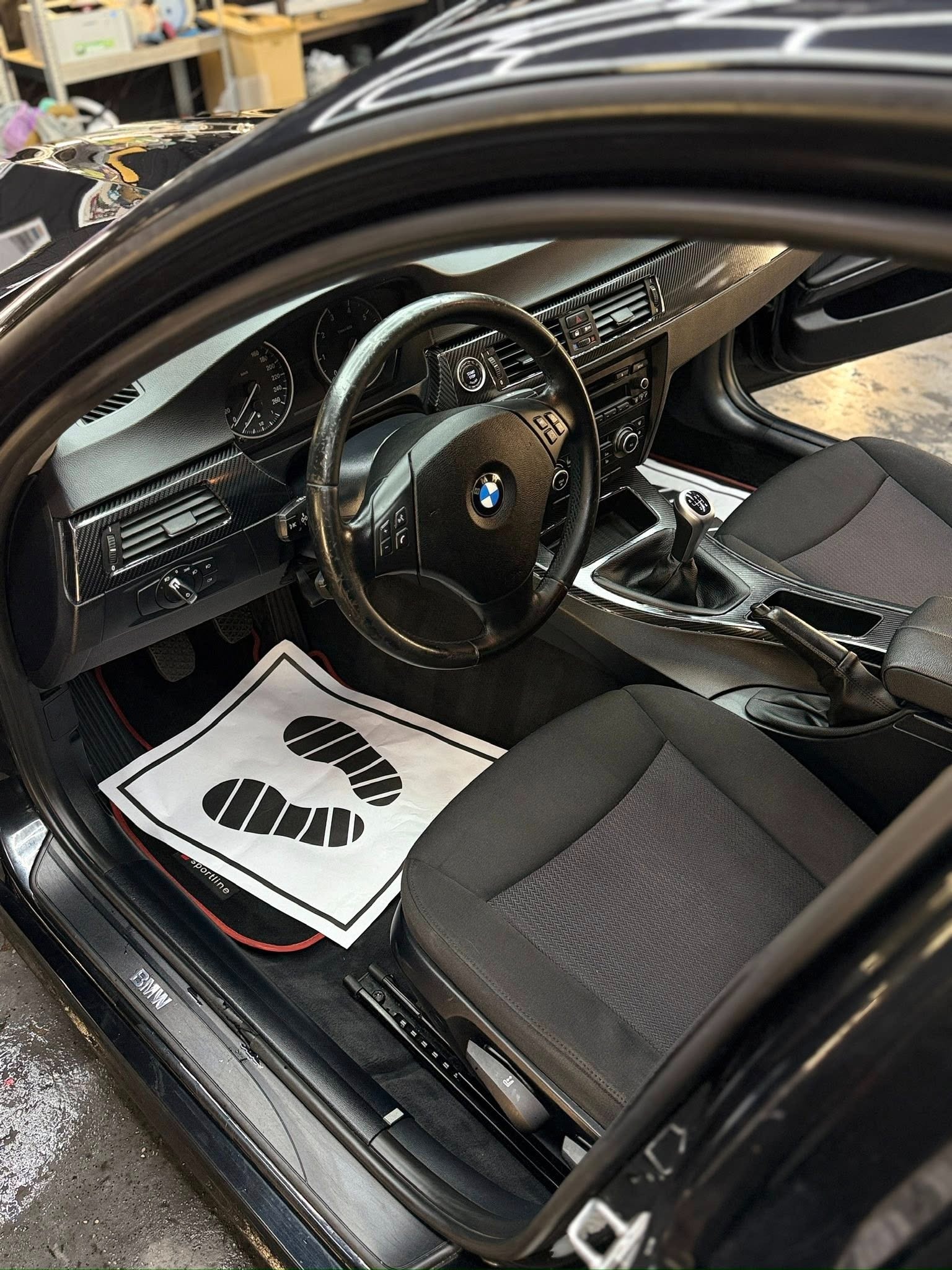 Wnętrze czarnego BMW E90 z karbonowymi akcentami na desce rozdzielczej i tunelu środkowym. Na podłodze leży papierowa mata z nadrukiem śladów stóp.