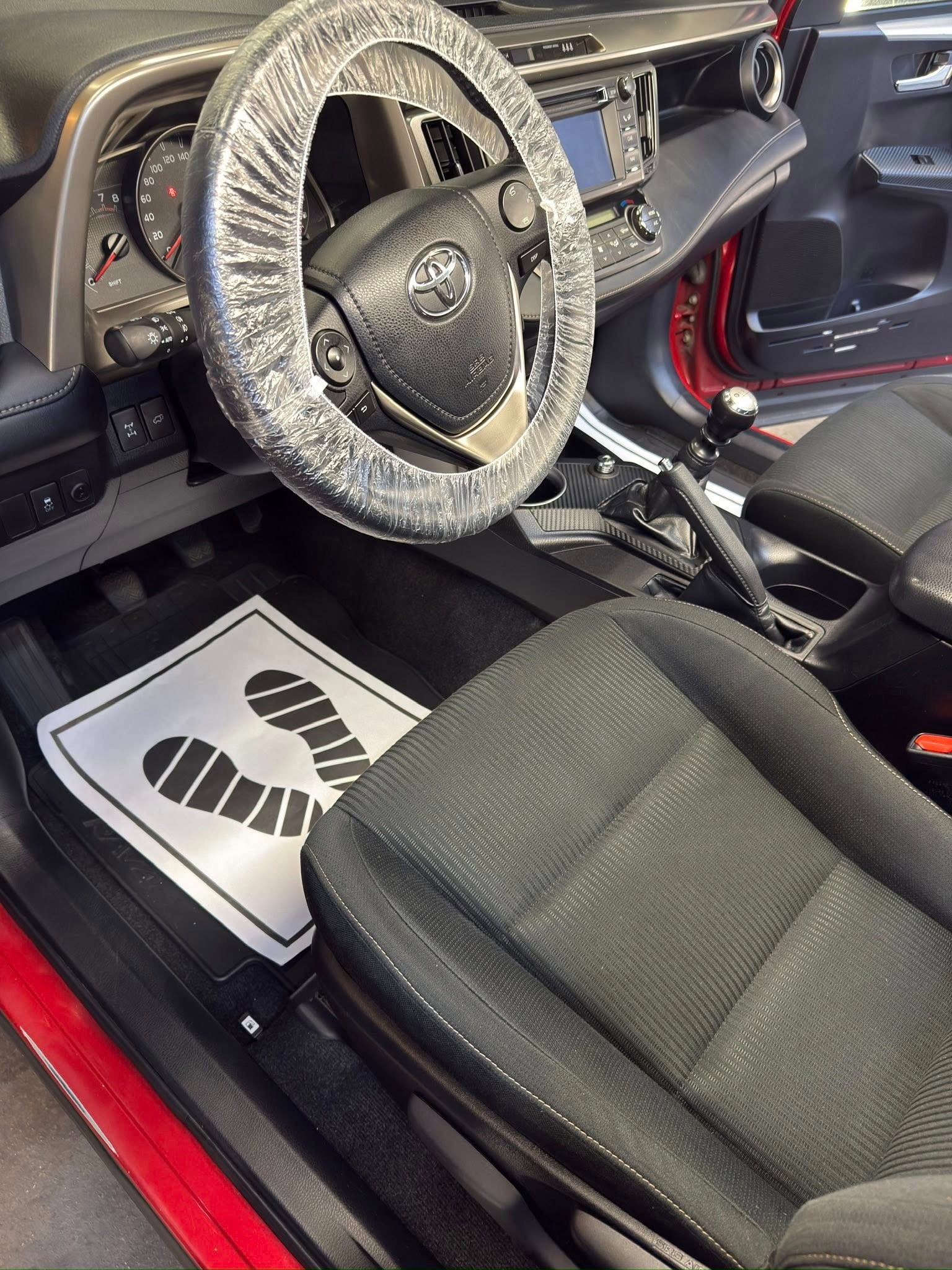 Wnętrze czerwonego auta z pokrowcem na kierownicy i papierową matą pod nogami. Widoczne czarne fotele i deska rozdzielcza. Przygotowanie do czyszczenia.